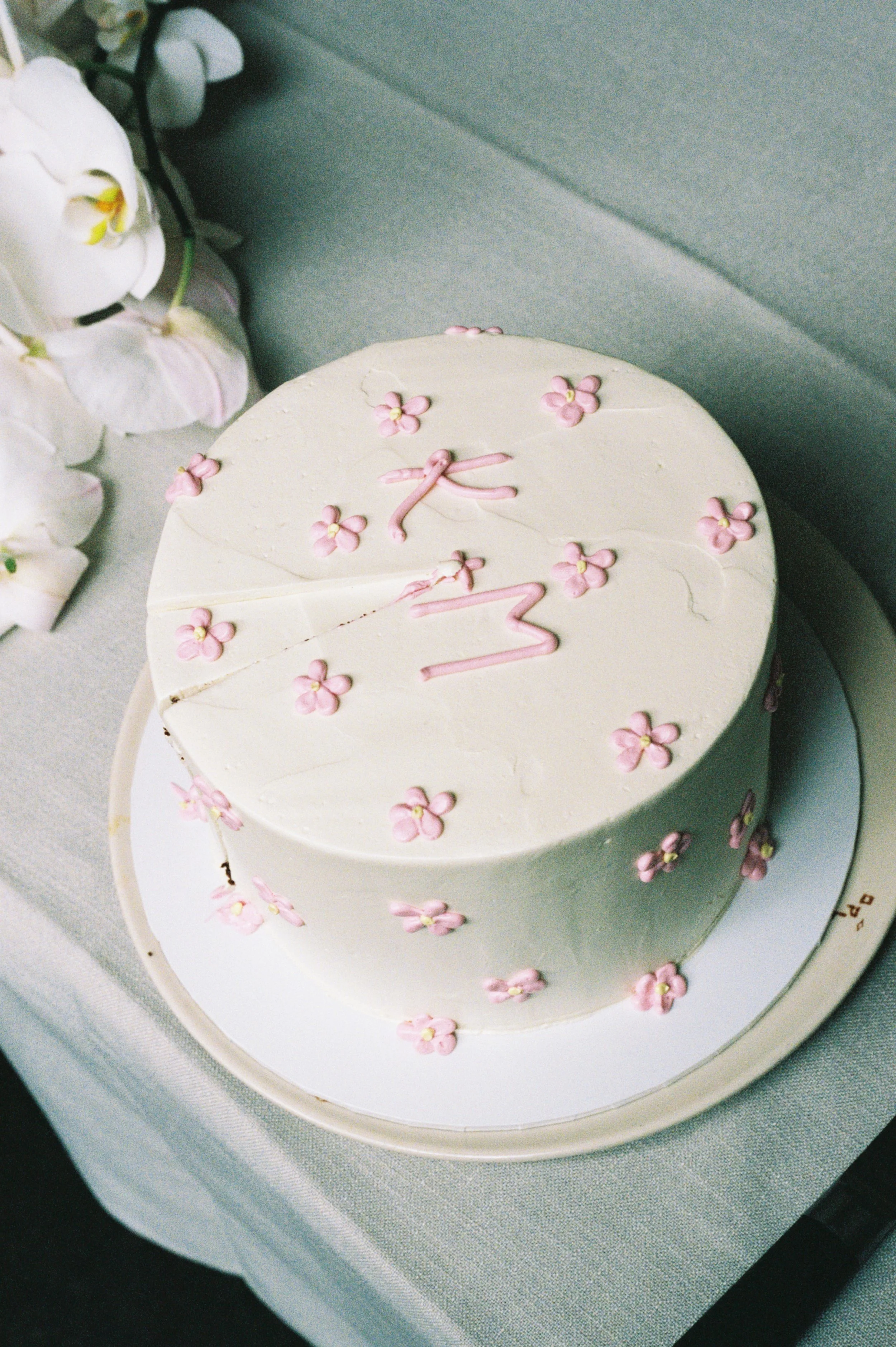 A round white cake decorated with pink flower-shaped candies and the words '1 M' written on top, placed on a white cake board.