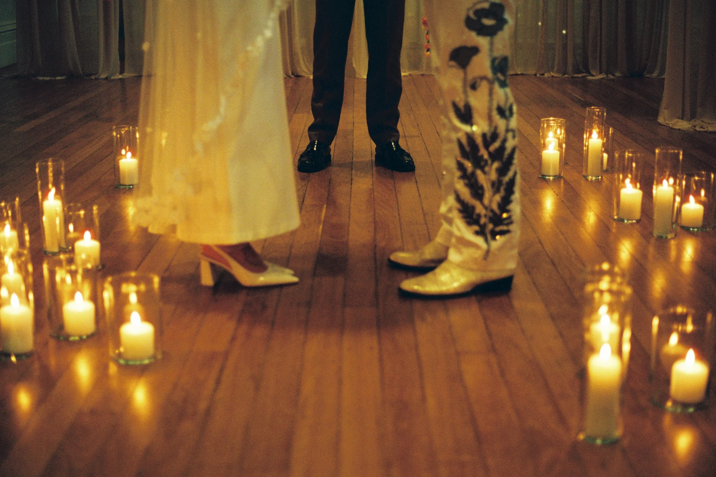 Close-up of a bride and groom's feet standing on a wooden floor, surrounded by lit candles, during a wedding ceremony.