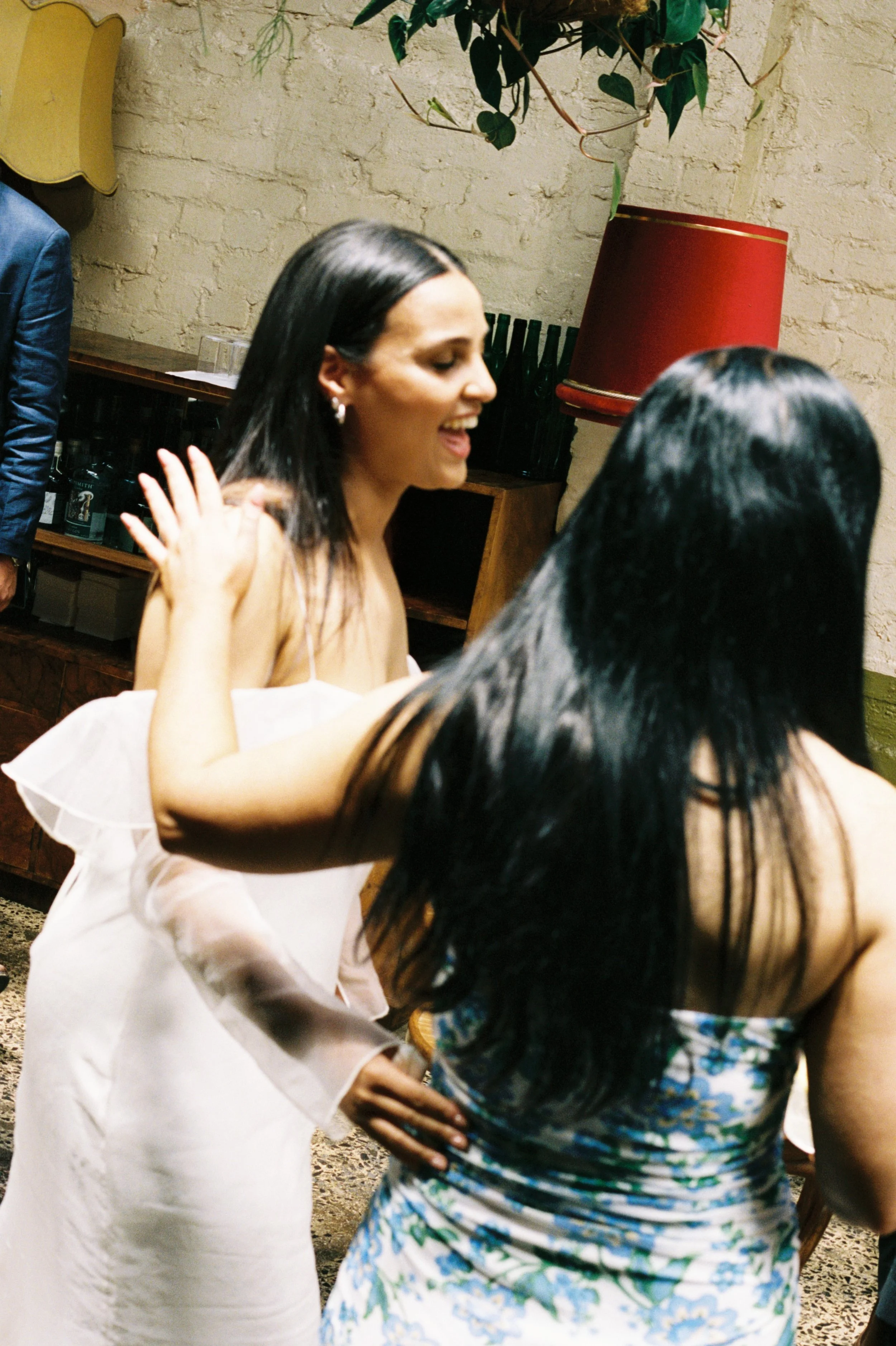 Two women dancing and smiling at a social gathering indoors, with a brick wall, a red lamp, and some greenery in the background.