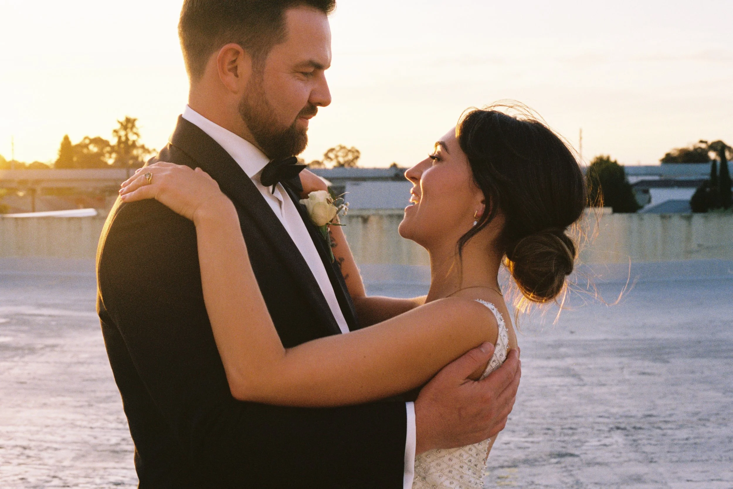 A newlywed couple dancing outdoors at sunset, with water in the background, the groom in a black tuxedo and the bride in a white wedding dress, smiling at each other.
