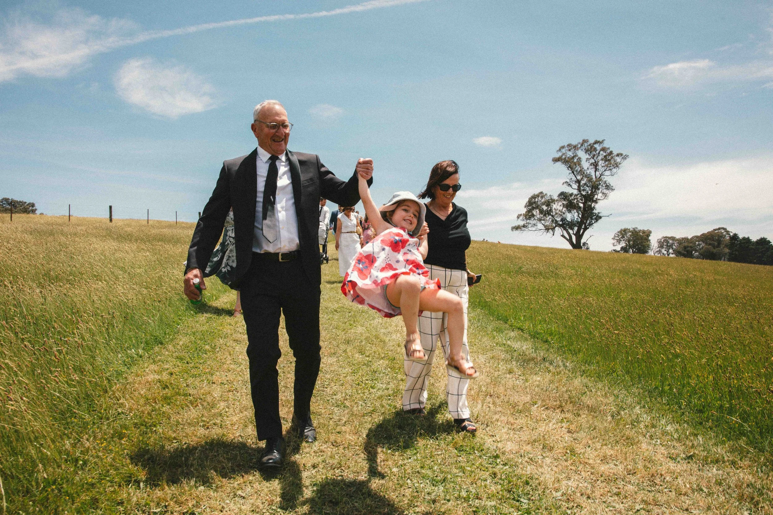 A young girl in a pink floral dress and sun hat being swung by two adults, a man in a black suit and a woman in patterned pants, on a grassy path in an open field with trees and a blue sky in the background.