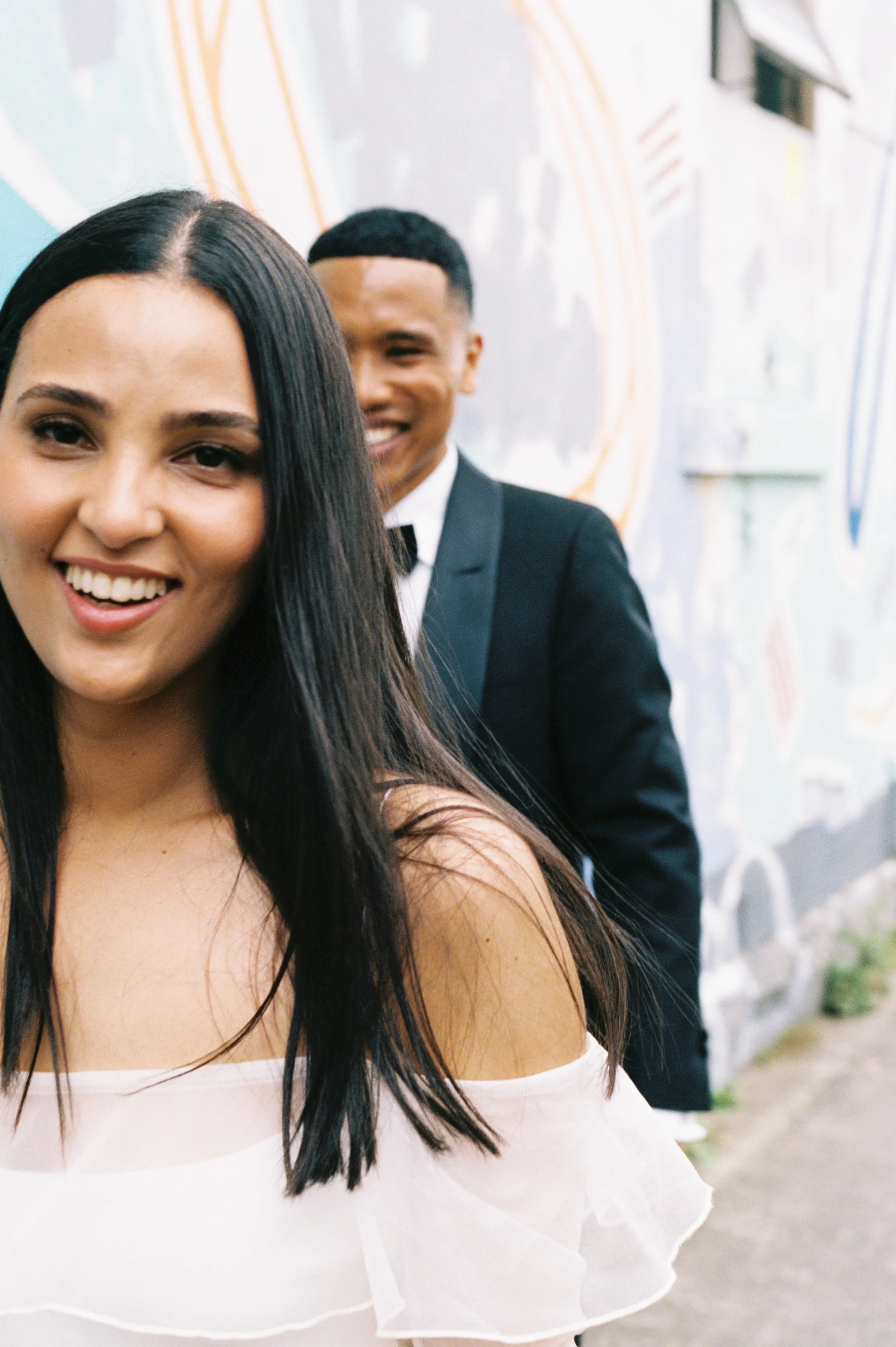 A woman with long dark hair and a white dress smiling in front of a man in a black tuxedo, standing in front of a colorful mural.
