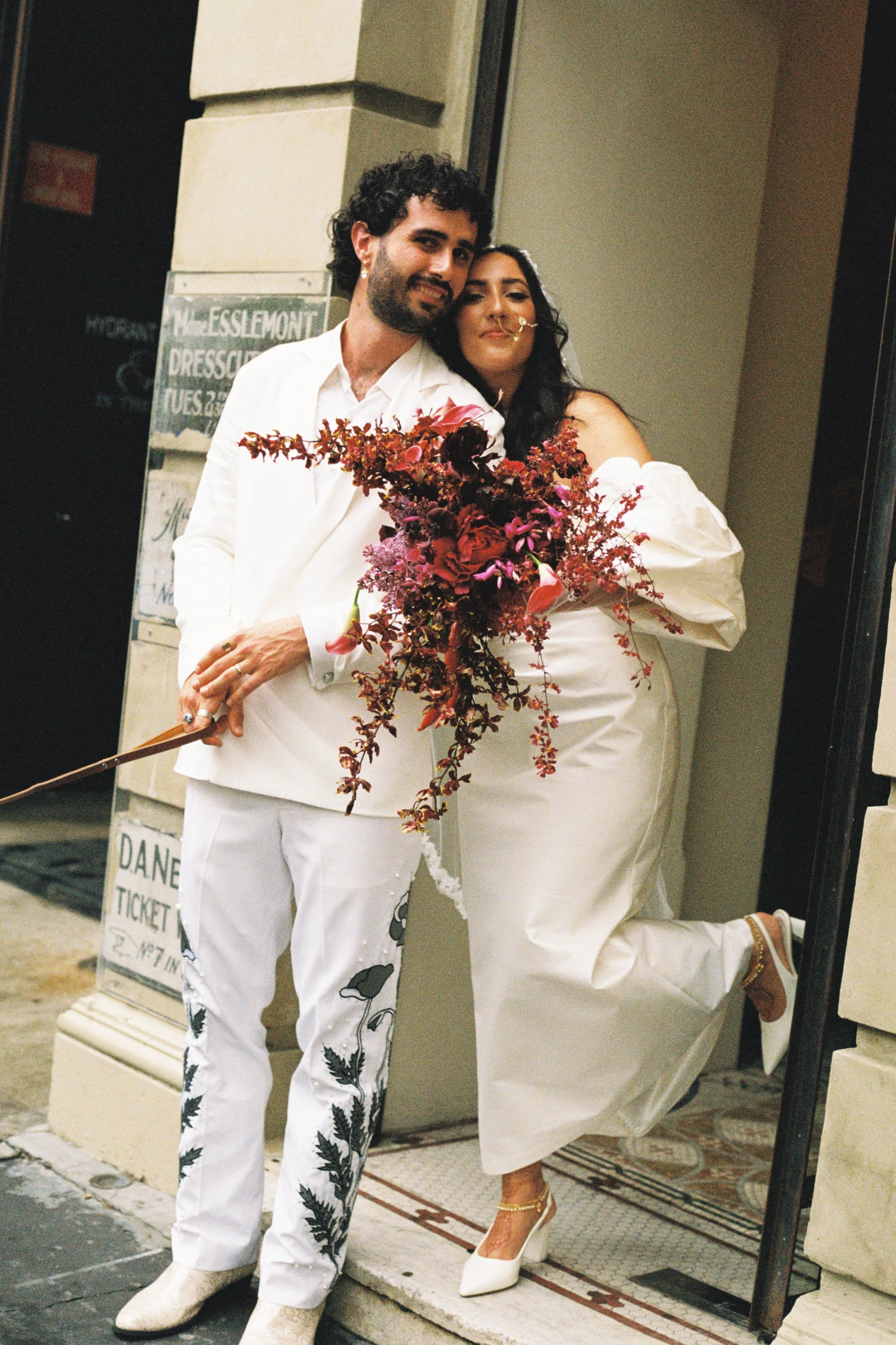 A couple in white outfits posing outside a building at night, holding a large bouquet of deep red and pink flowers.