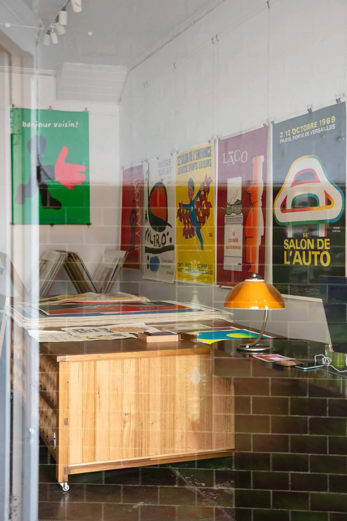 Interior view of a room seen through a window, showing posters on the wall, a wooden table with papers, and a yellow desk lamp. The posters include colorful designs and text in French.