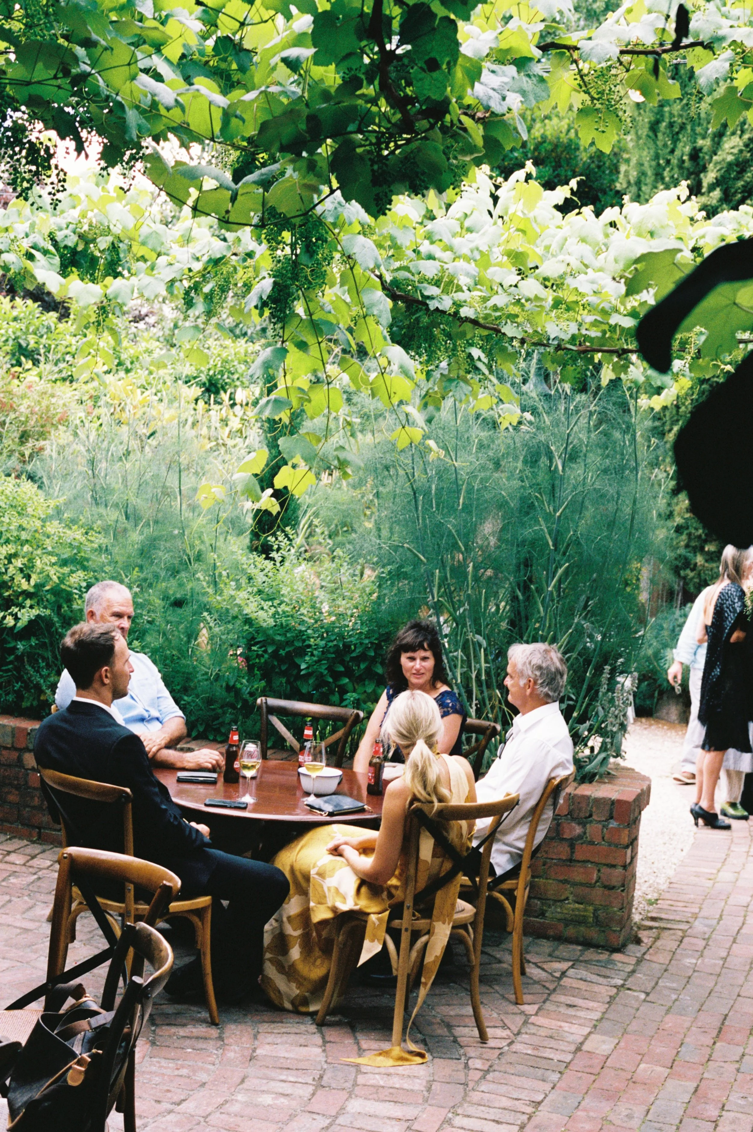 Group of six people sitting around a wooden table outdoors in a lush green garden, engaged in conversation at a social gathering.