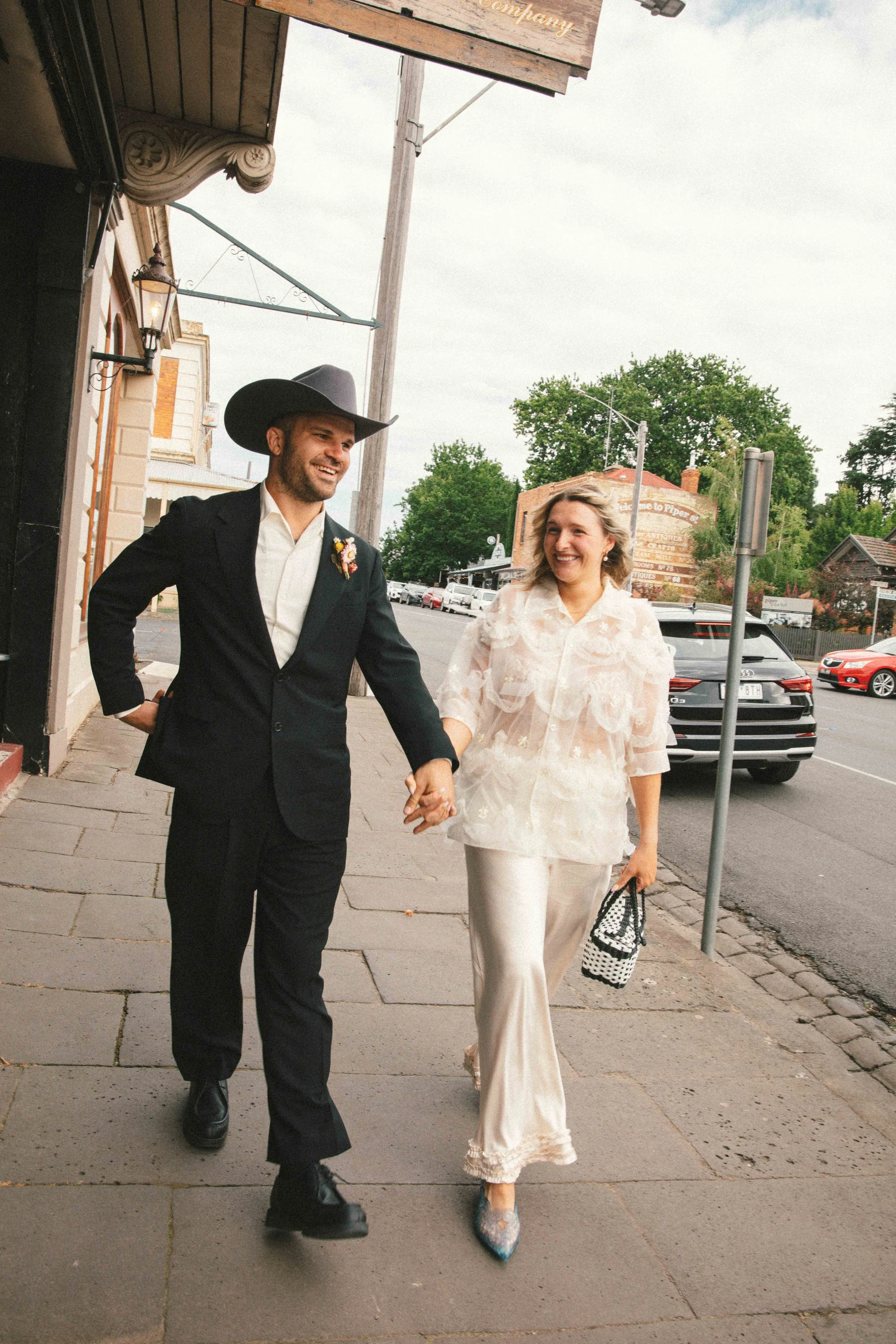 A smiling man in a black suit and cowboy hat holding hands with a smiling woman in an elegant cream outfit walking on a sidewalk in a small town.