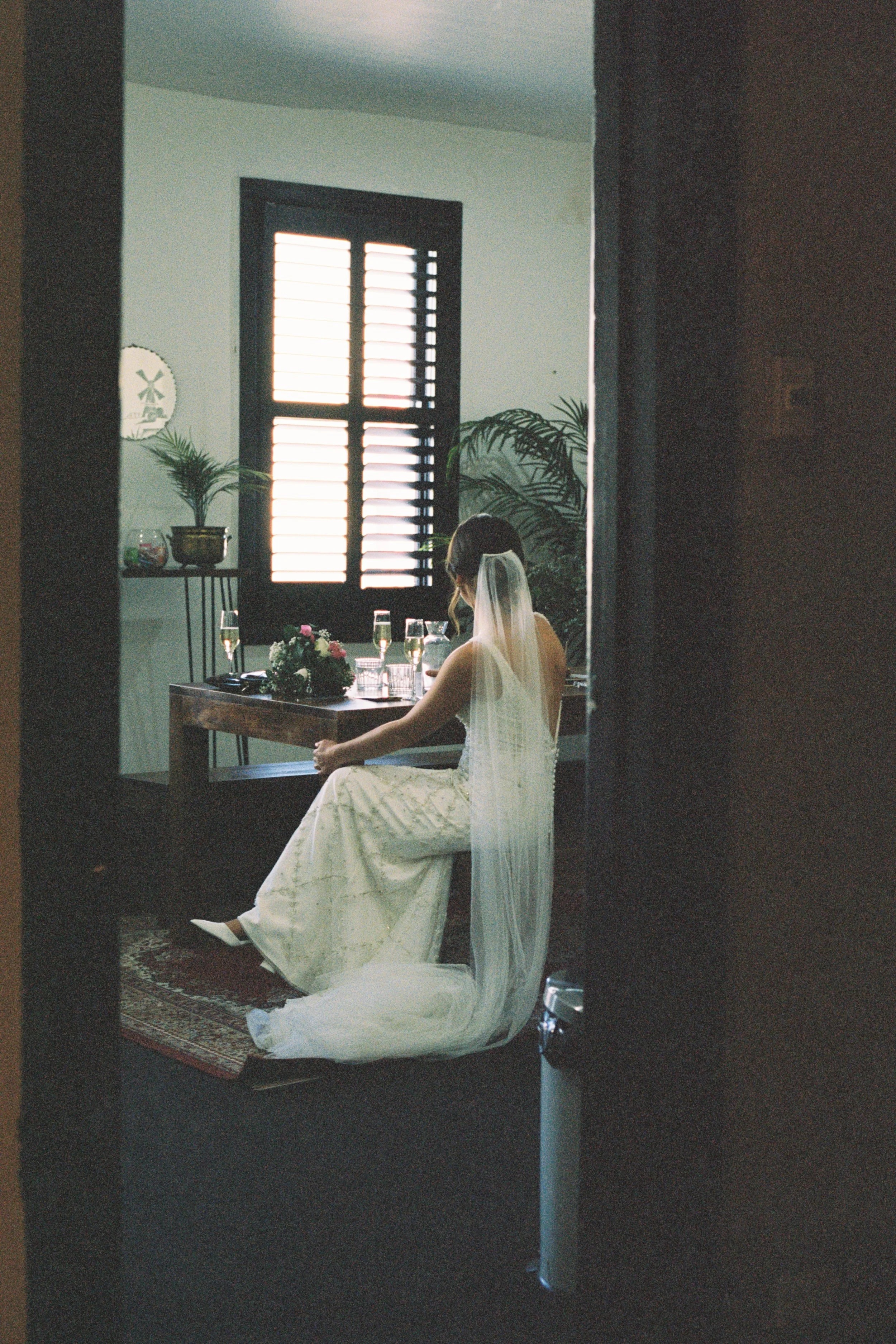 A bride in a wedding dress and veil sitting at a table with champagne glasses and a floral centerpiece, in a room with a window and plants in the background.
