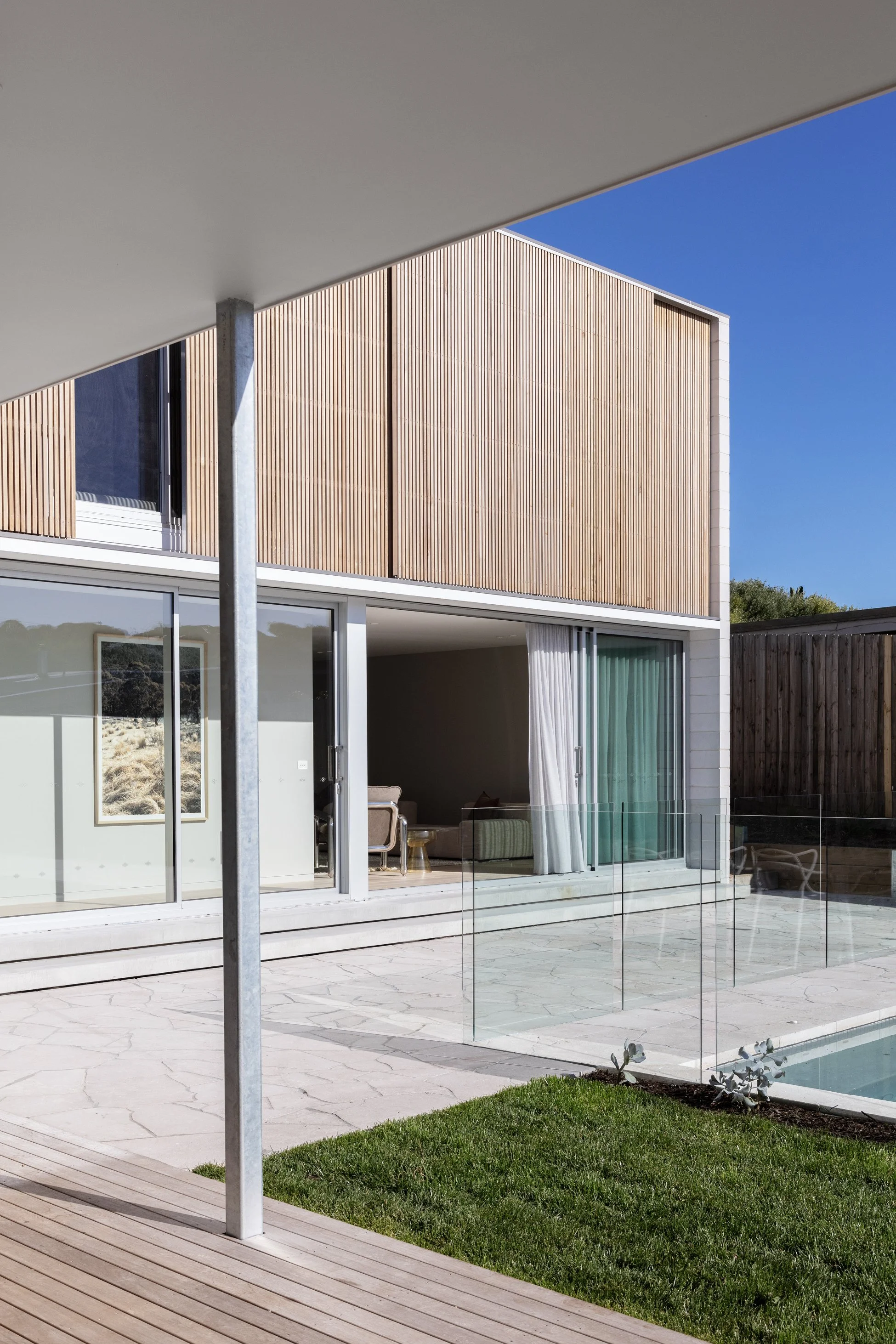 Modern two-story house with wood-paneled exterior, sliding glass doors, outdoor patio, swimming pool, and green lawn under a clear blue sky.