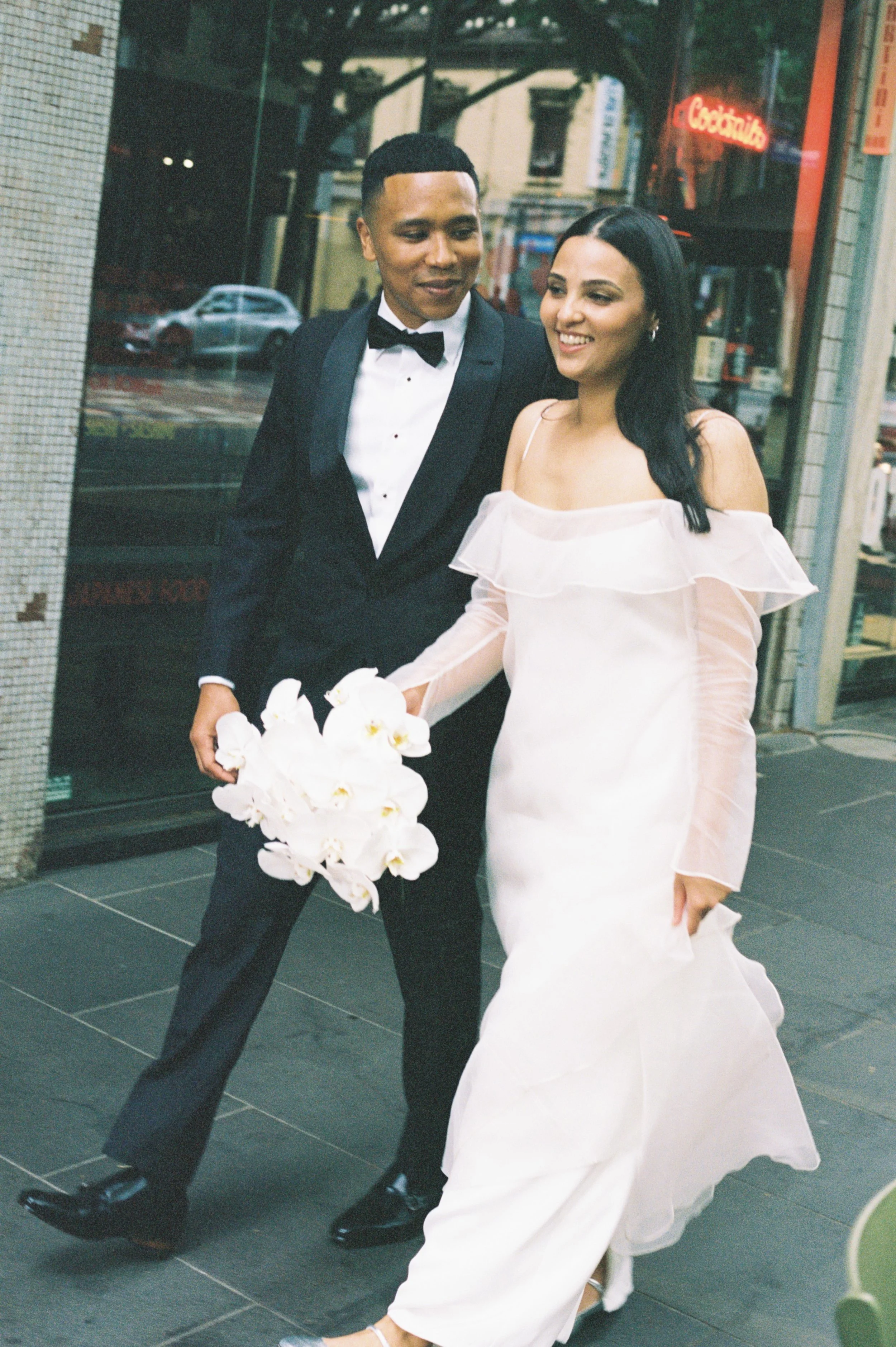 A newlywed couple walking on a city sidewalk, the groom in a suit with a bow tie holding a bouquet of white orchids, the bride in a white off-shoulder dress smiling.