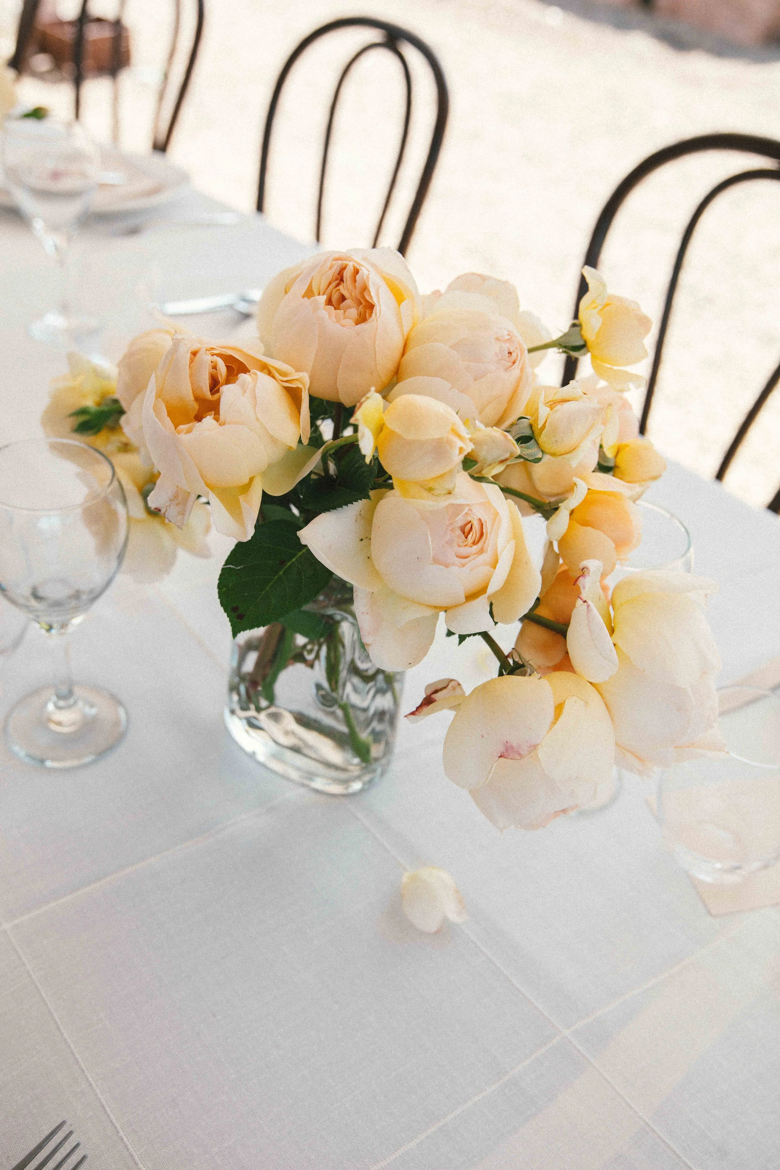 A table decorated with a vase of pale peach-colored peonies and rose flowers, surrounded by empty wine glasses and silverware, set for a formal event.