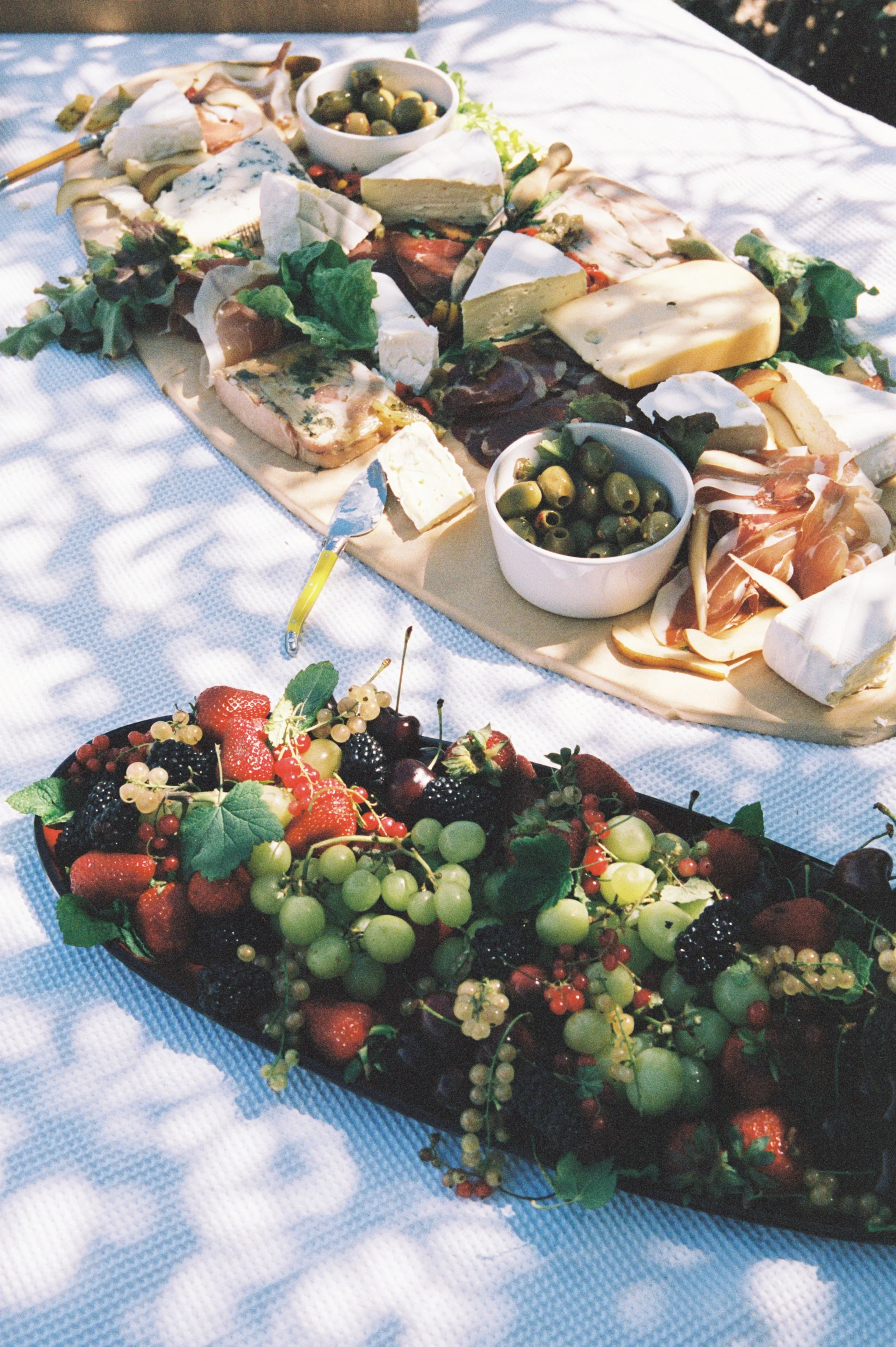 A picnic table with a cheese platter, bowls of green olives, and a large tray of mixed fresh berries, grapes, strawberries, blackberries, and red currants.