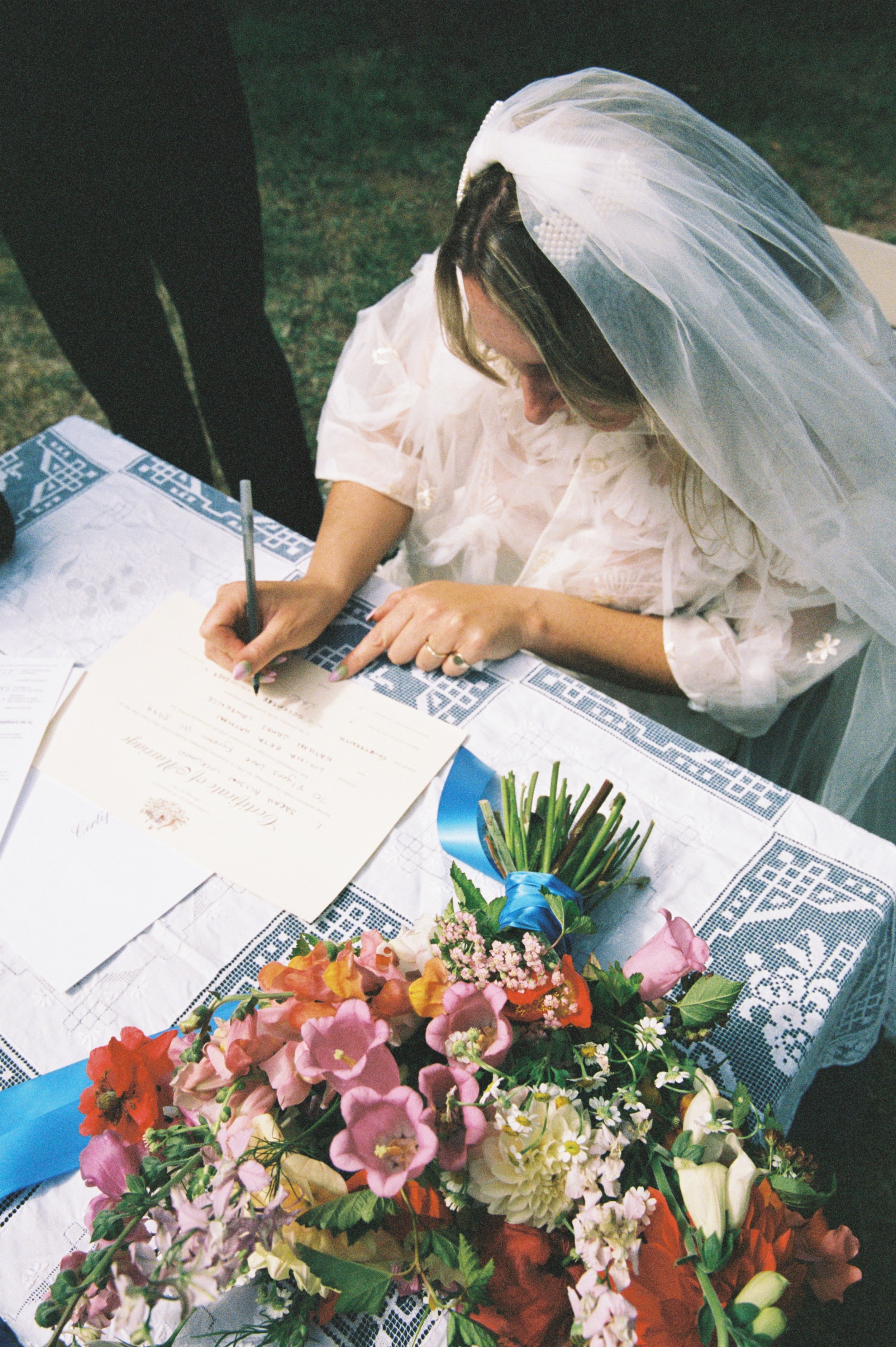 A woman wearing a bridal veil signs a document at a table covered with a lace cloth, with a bouquet of colorful flowers in the foreground.