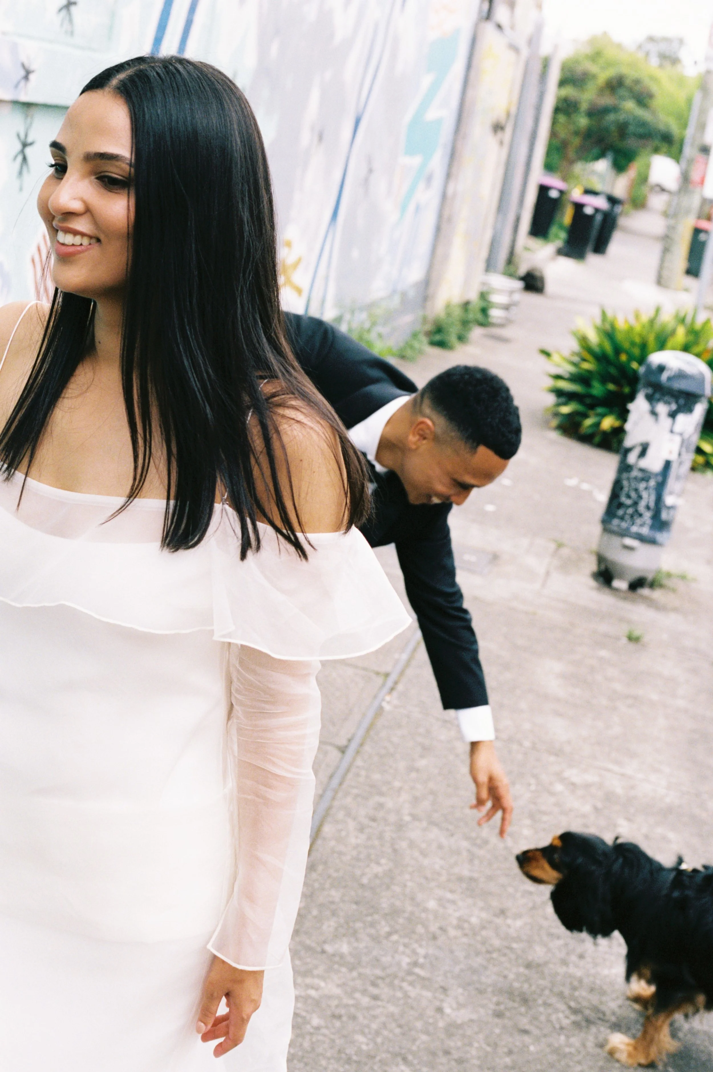 A woman in a white dress smiling while standing on a sidewalk, with a man in a suit reaching down to pet a black and tan dog nearby.