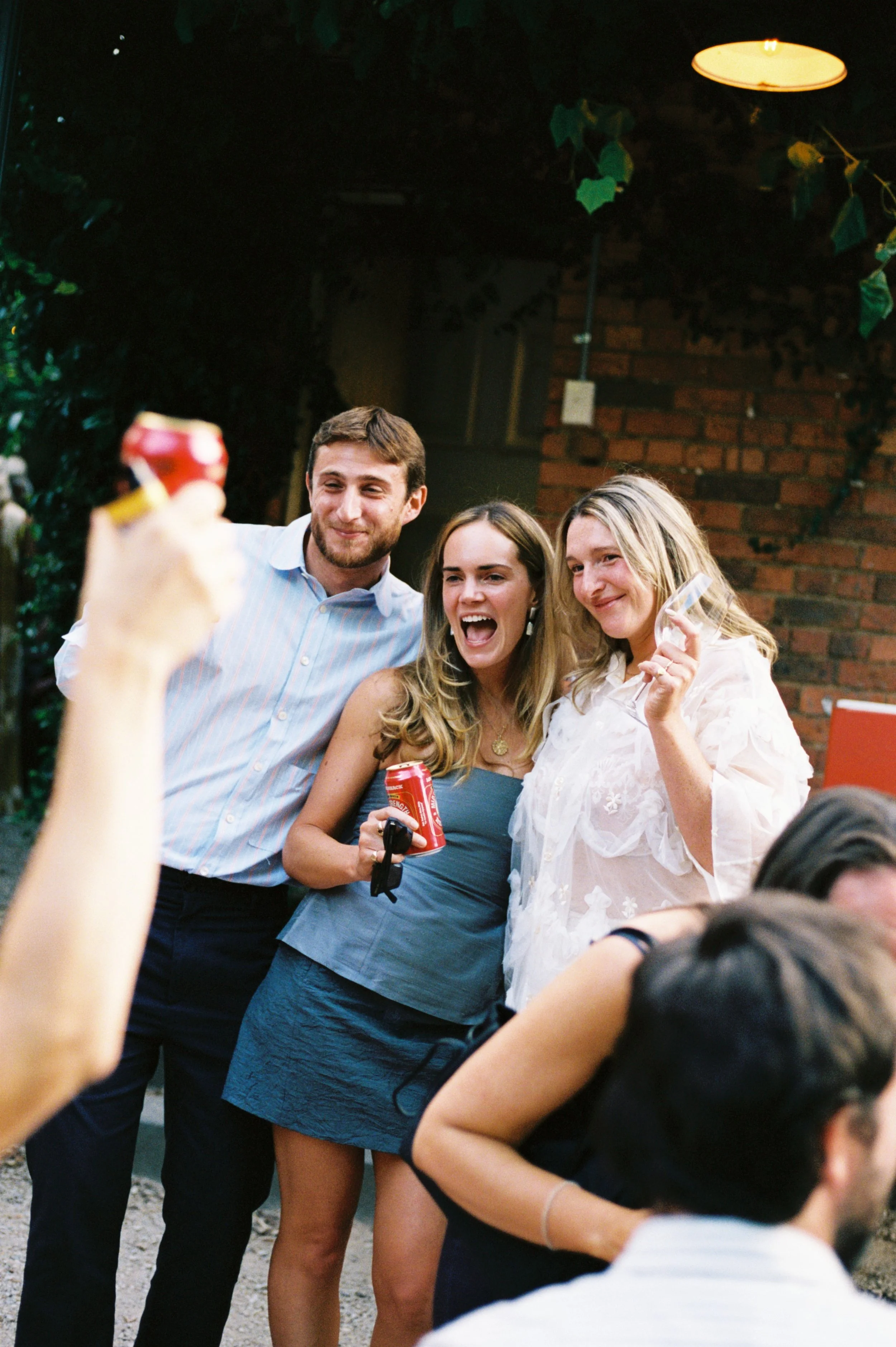 Three friends, two women and one man, standing together smiling and posing for a photo at a social gathering outdoors at night. One woman is holding a can of soda, another woman holds a wine glass, and the man is smiling. They are close to each other