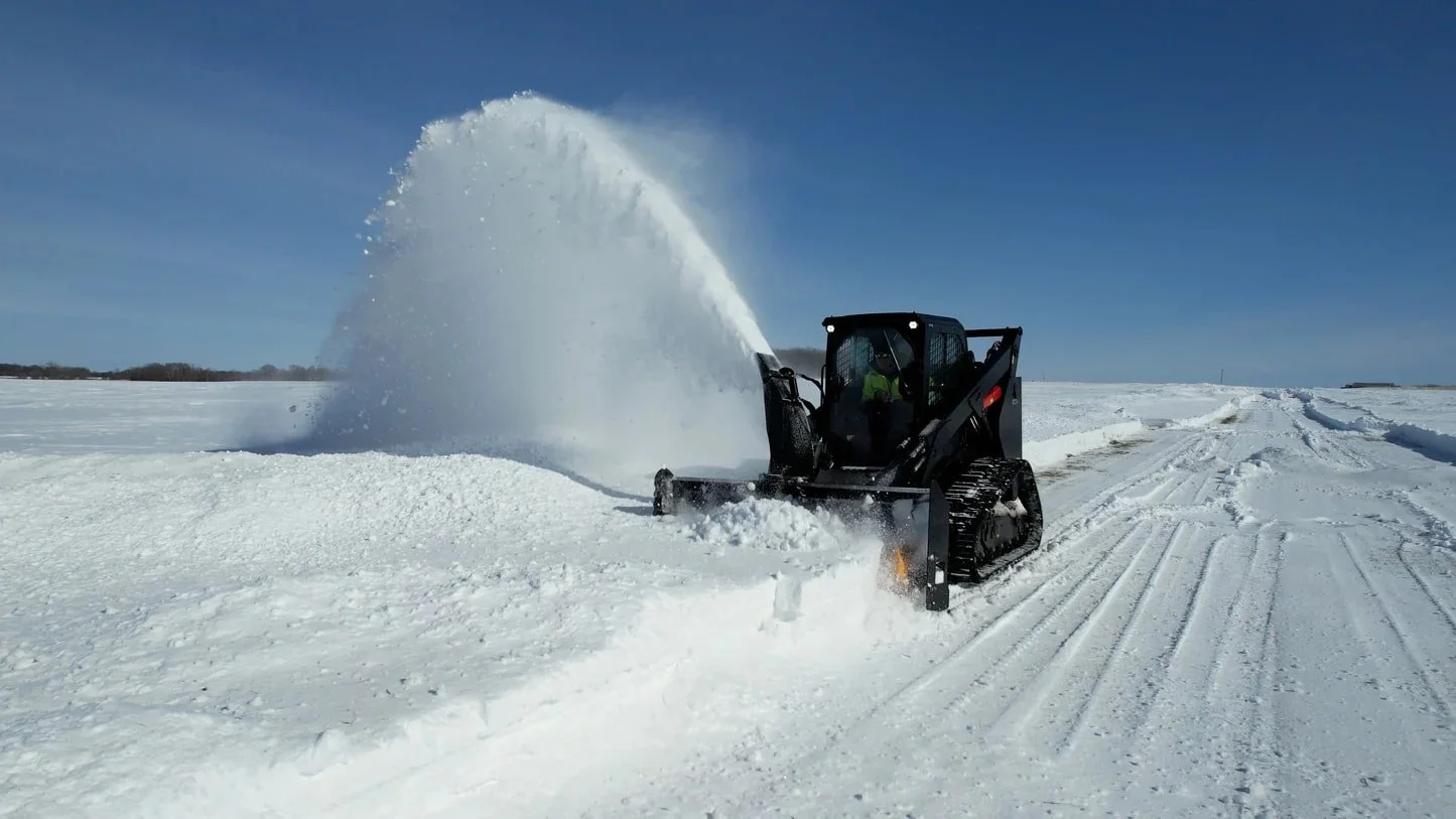 A black Skid Steer using the Quick Attach Summit Series snowblower attachment on a snow-covered road in a snowy landscape during daytime.