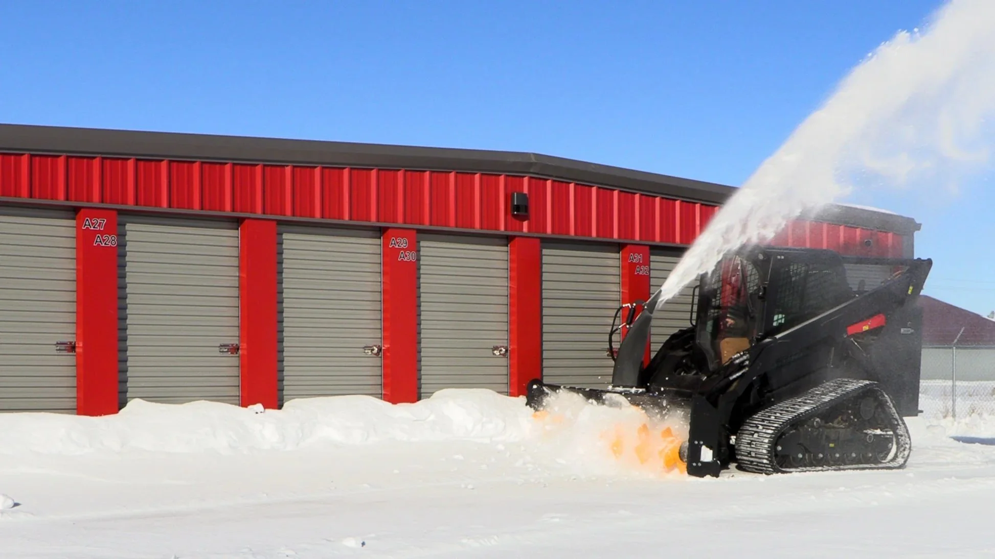 A skid steer with the Quick Attach Summit Series snowblower machine clearing snow in front of a red storage unit building on a sunny day.