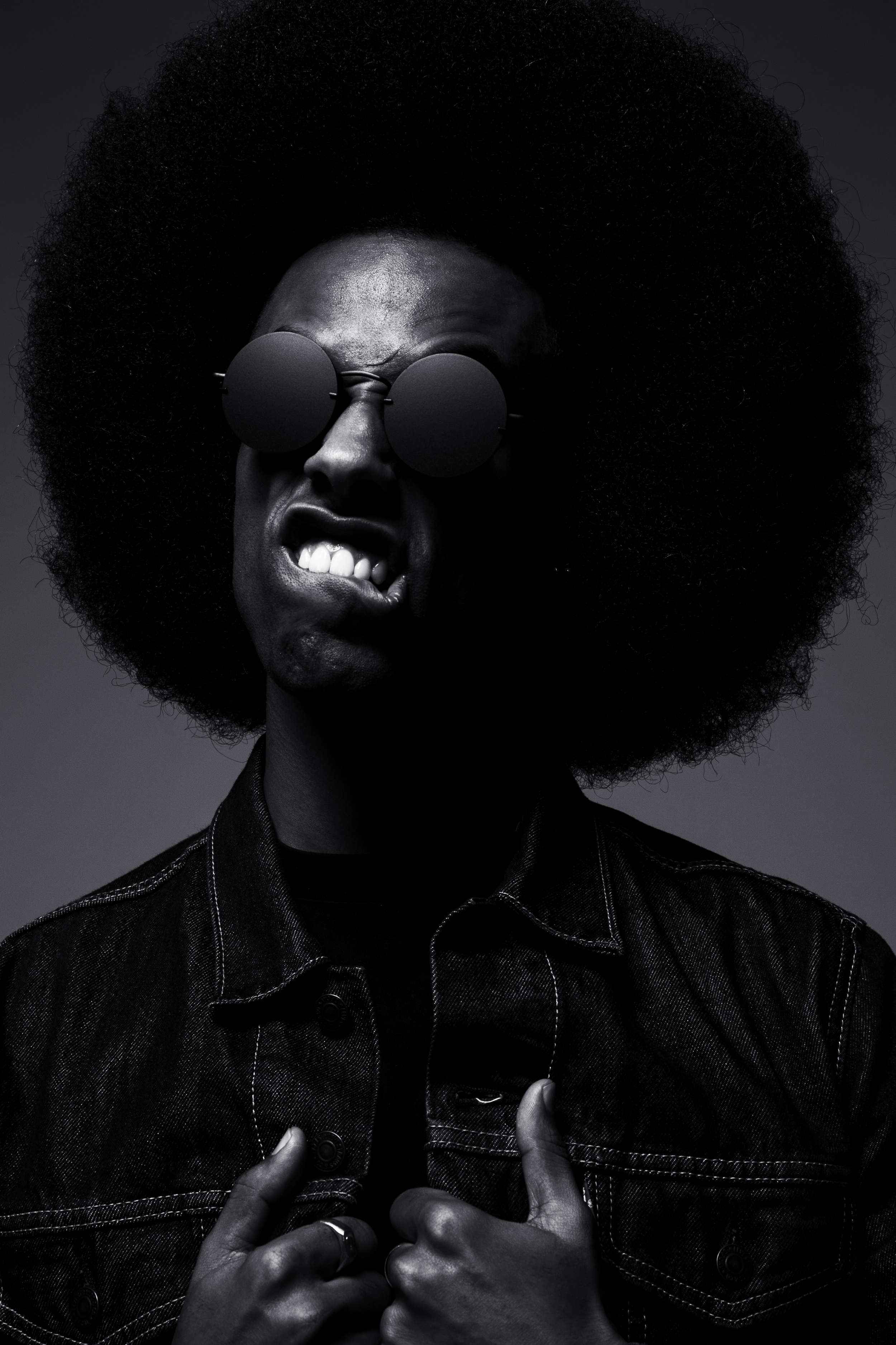 Portrait of a person with an afro hairstyle, wearing round sunglasses and a denim jacket, making an expressive facial expression.