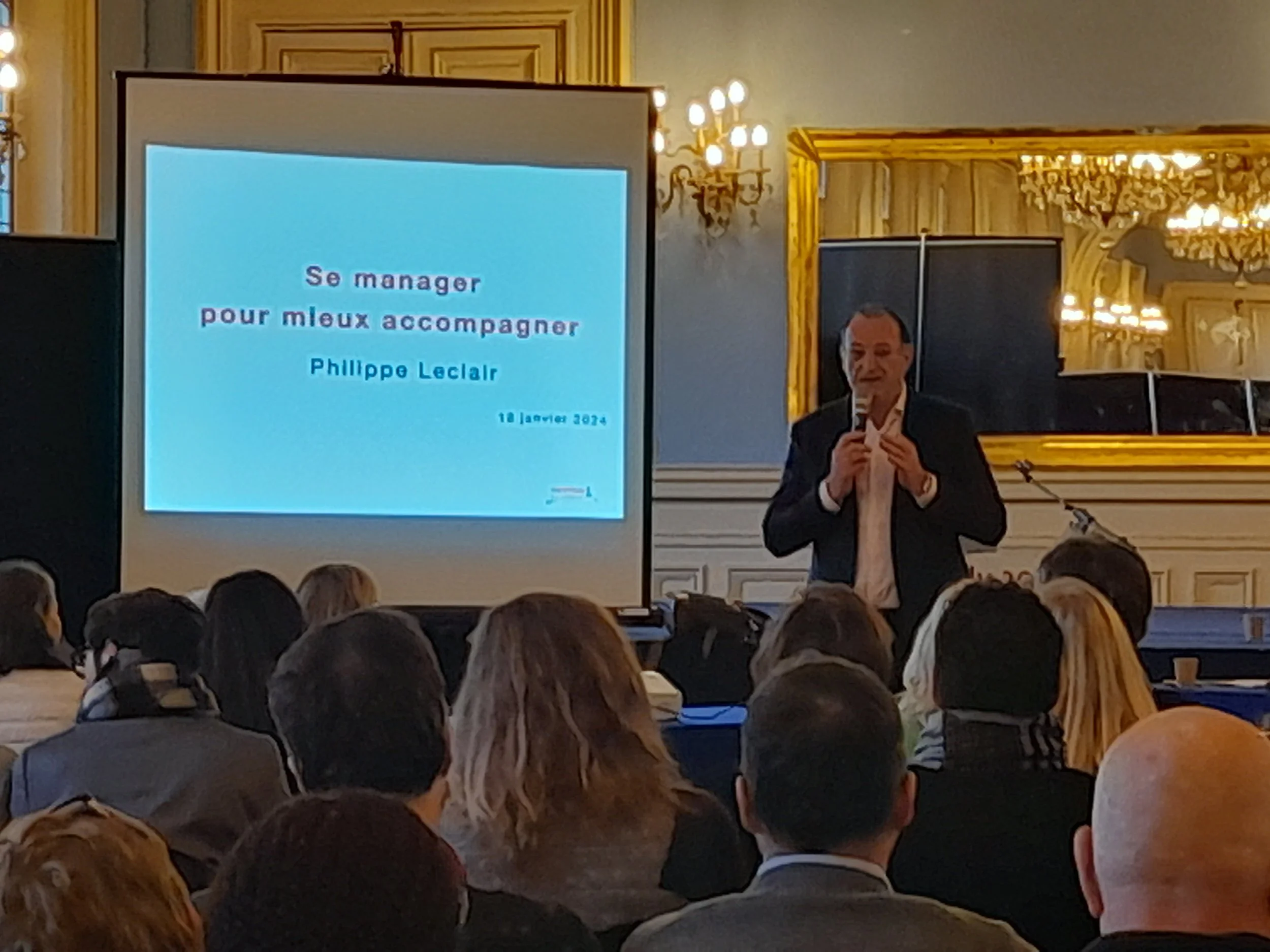 Une conférence avec un homme parlant dans un micro, un écran affichant le titre « Se manger pour mieux accompagner » par Philippe Leclaire, dans une salle élégante avec des lustres en cristal et des murs en bois ornés.