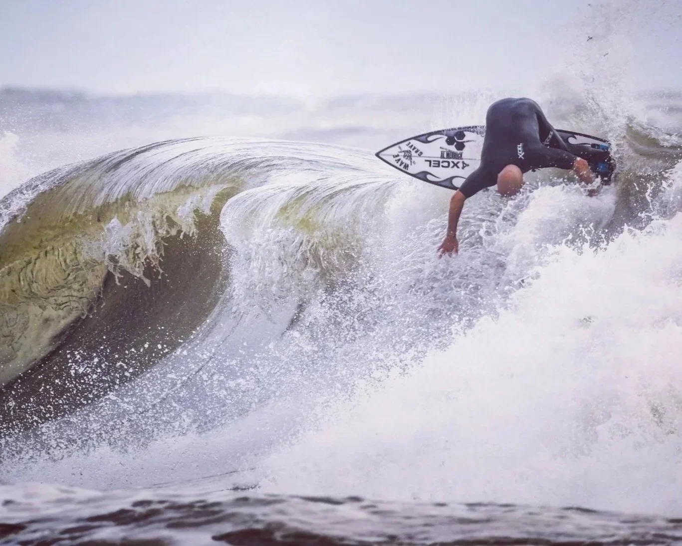 A person surfing on a wave in rough water, wearing a black wetsuit and hunched over on a surfboard.