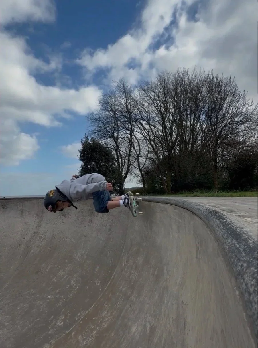 A young boy in a gray hoodie, helmet, and shorts is hanging upside down on the edge of a concrete skatepark bowl with trees and a partly cloudy sky in the background.