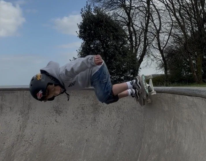 A young boy wearing a helmet, gray hoodie, shorts, and rollerblades, hanging upside down over the edge of a concrete skate park bowl, with trees and a cloudy sky in the background.