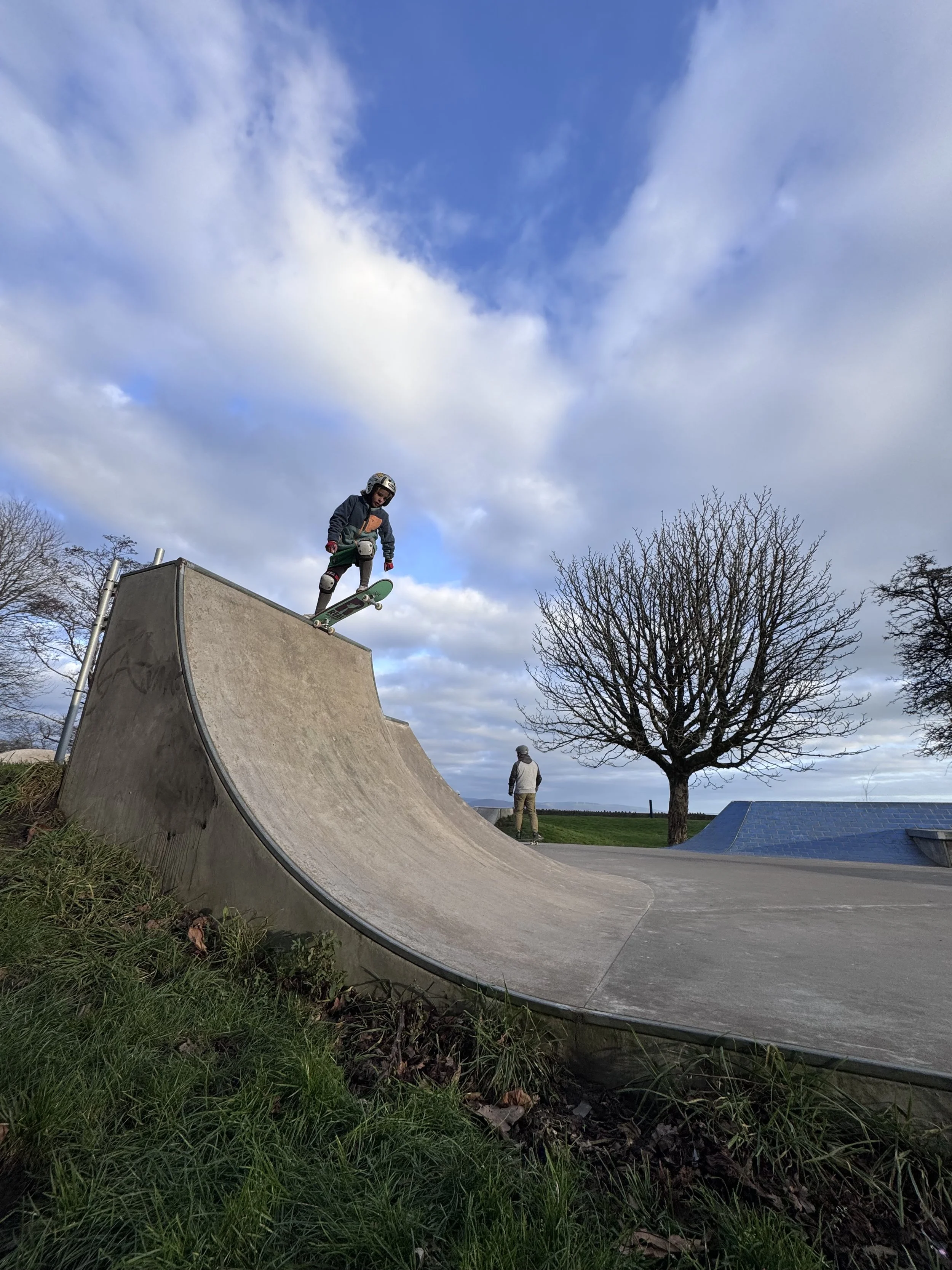 A person riding a skateboard on a ramp at a skatepark with another person and a leafless tree in the background under a partly cloudy sky.