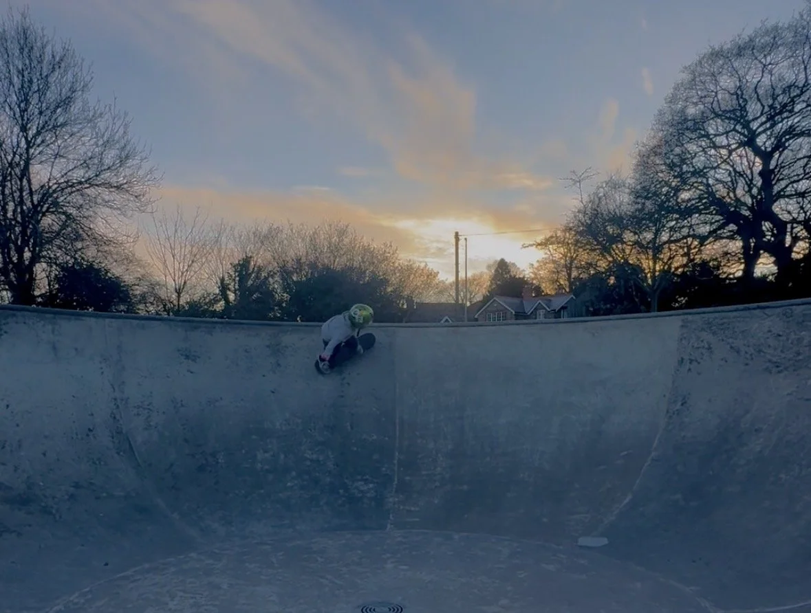 A person wearing a helmet skateboarding in a skate bowl at sunset with bare trees and houses in the background.