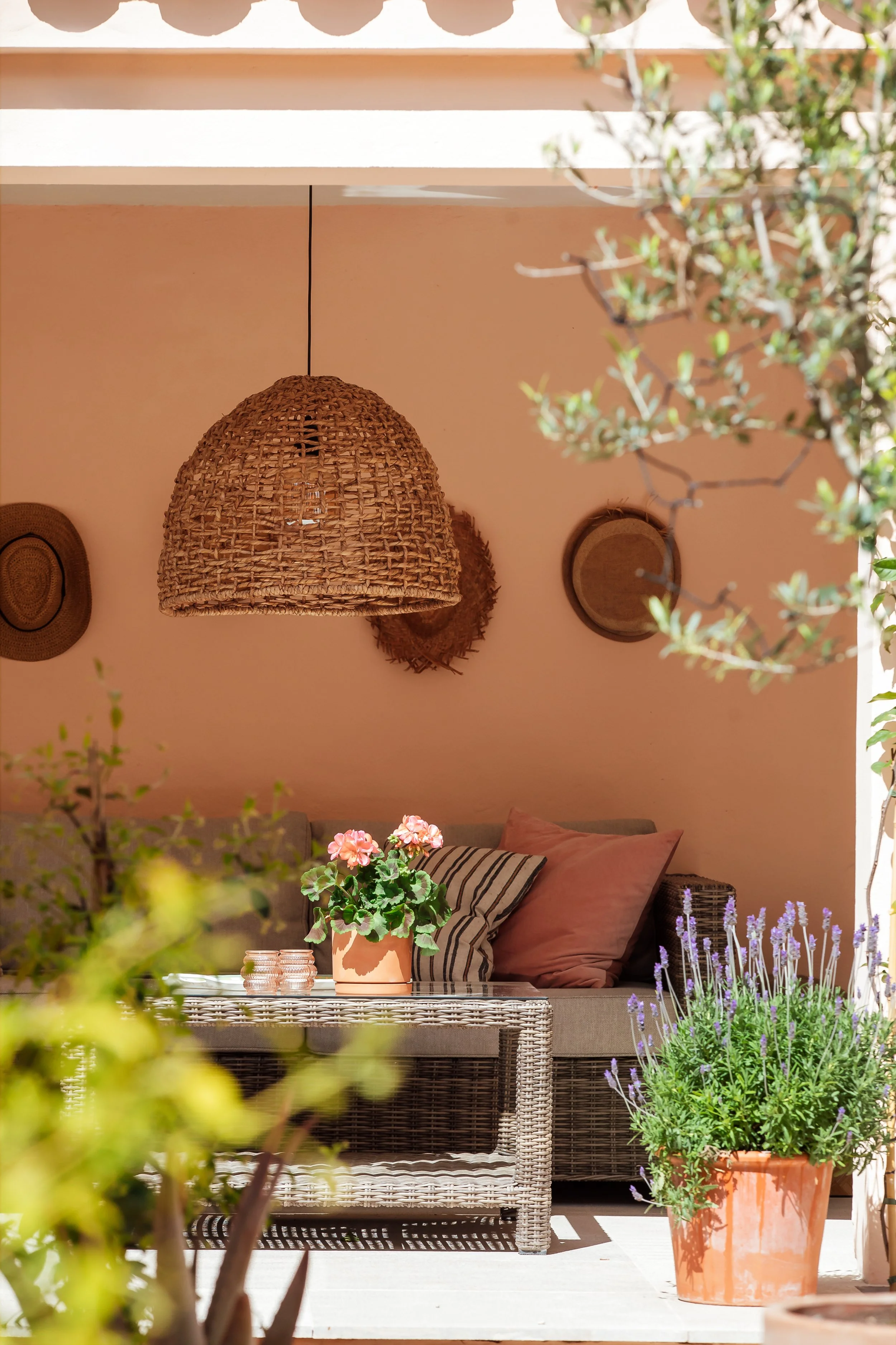 An outdoor patio scene with potted plants, floral cushions, a wicker coffee table, and a woven pendant light hanging above.