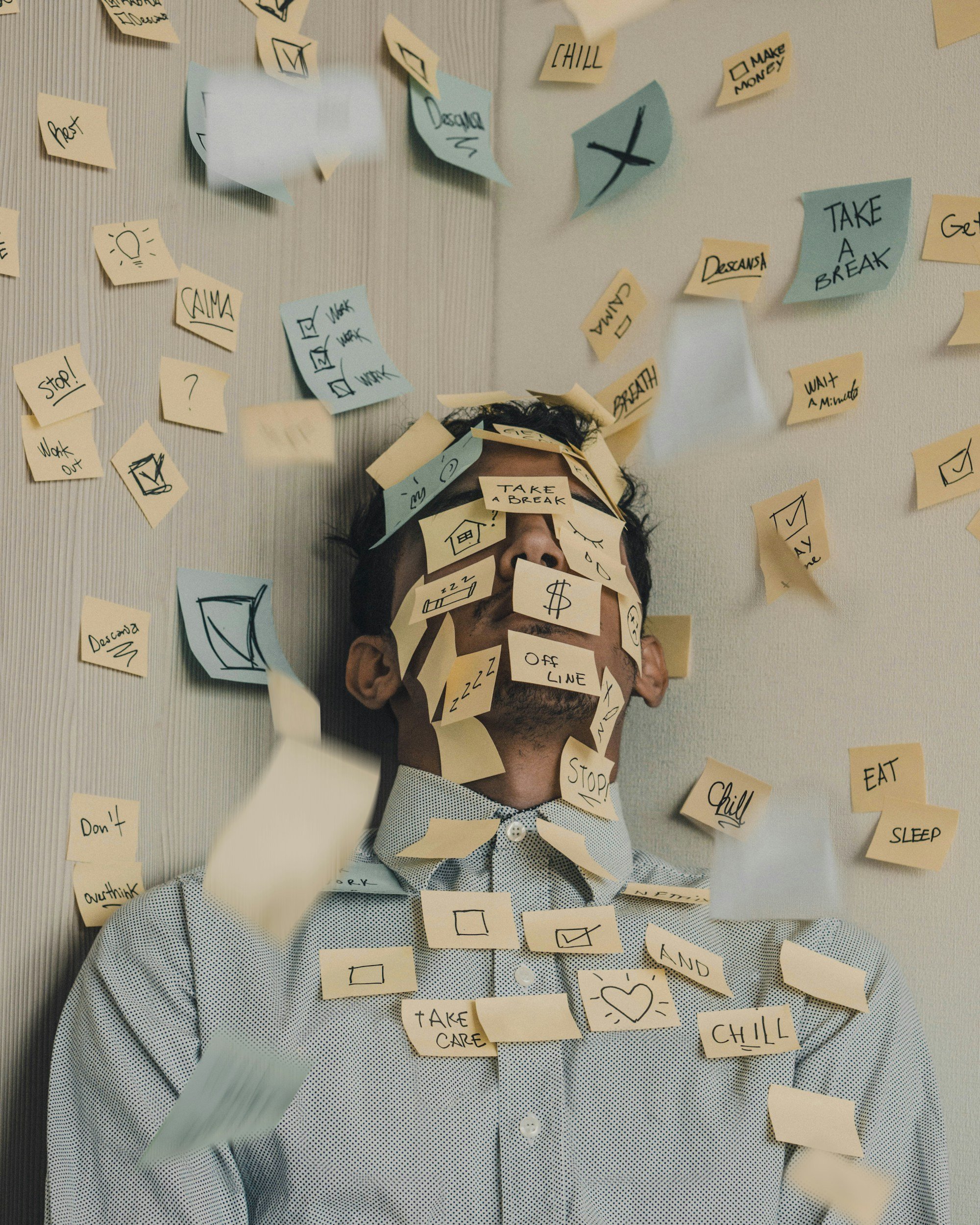En person med kort, mörkt hår som bär en ljusblå och vit prickig skjorta har många gula och blå post-it-lappar på ansiktet, halsen och kläderna. Lapparna innehåller olika handskrivna ord, fraser, kryssrutor, symboler och teckningar, till exempel ”TAKE A BREAK”, ”Chill”, ”Sleep”, ”Stop!”, ”Don't”, ”Outlook”, ”$$” och en glödlampsikon.