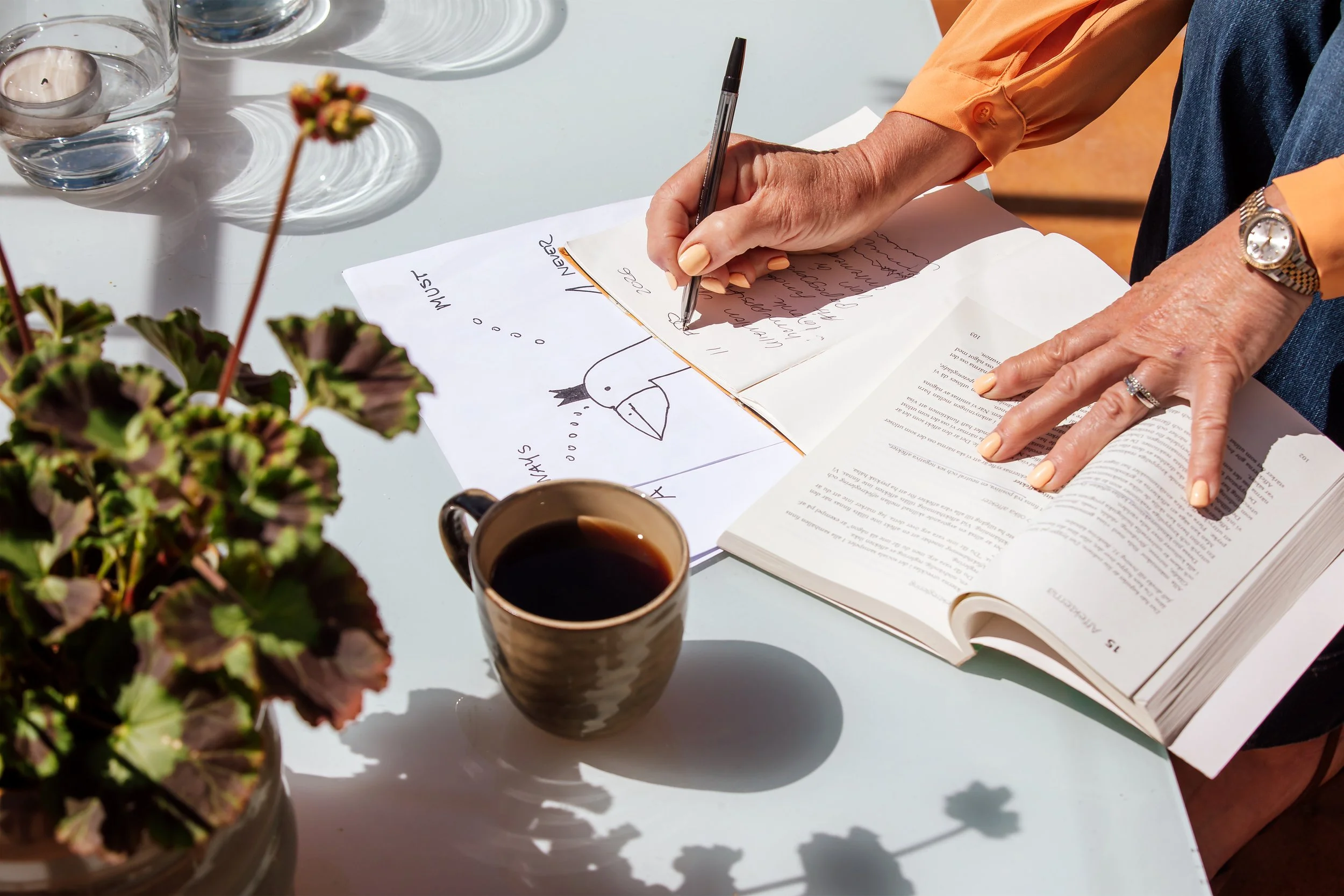 A person writes in a notebook with a pen, surrounded by a book, a cup of coffee, and glasses on a white table with sunlight casting shadows.