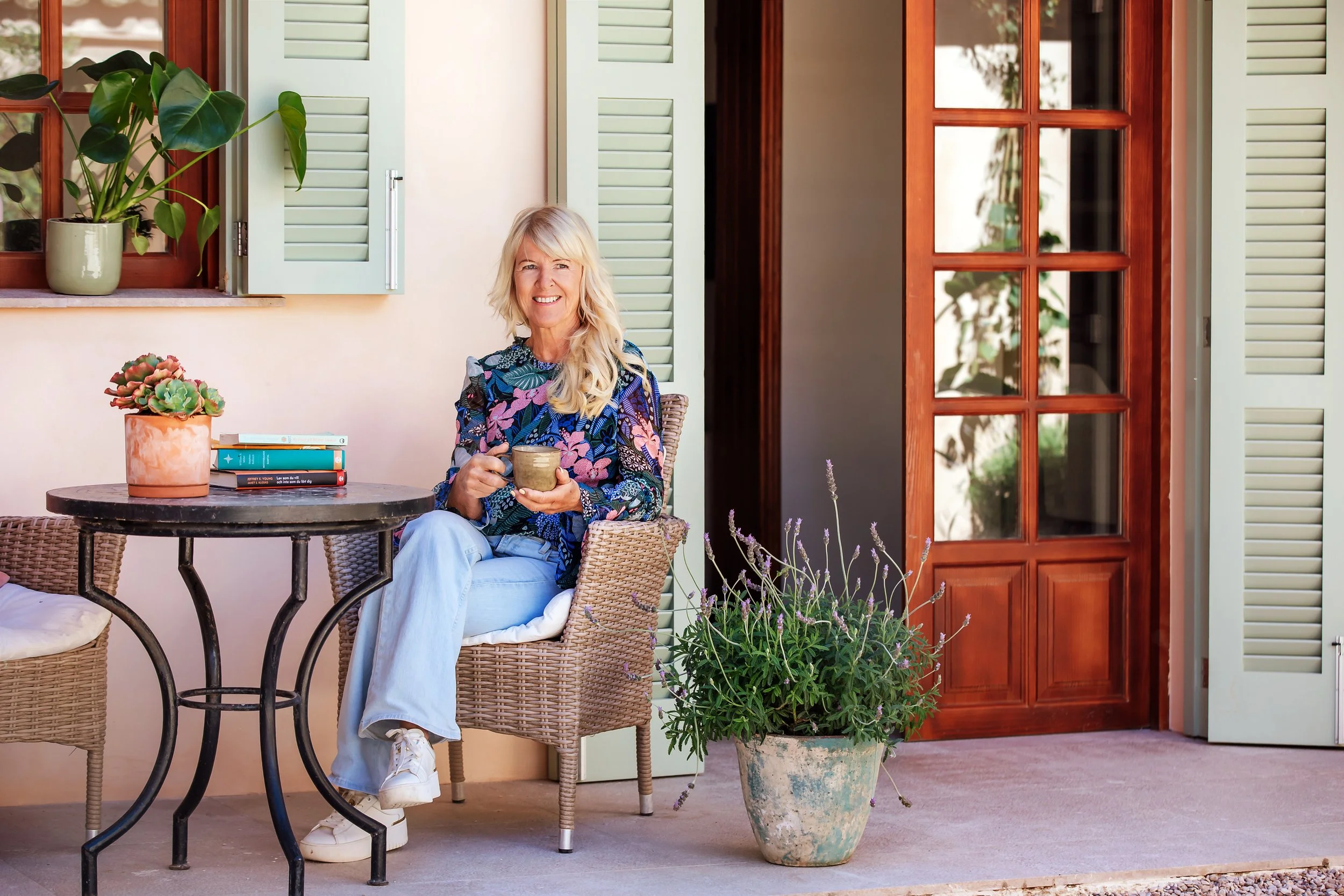 A woman sitting outside on a porch holding a mug, surrounded by potted plants and flowers, with open window shutters and a door in the background.