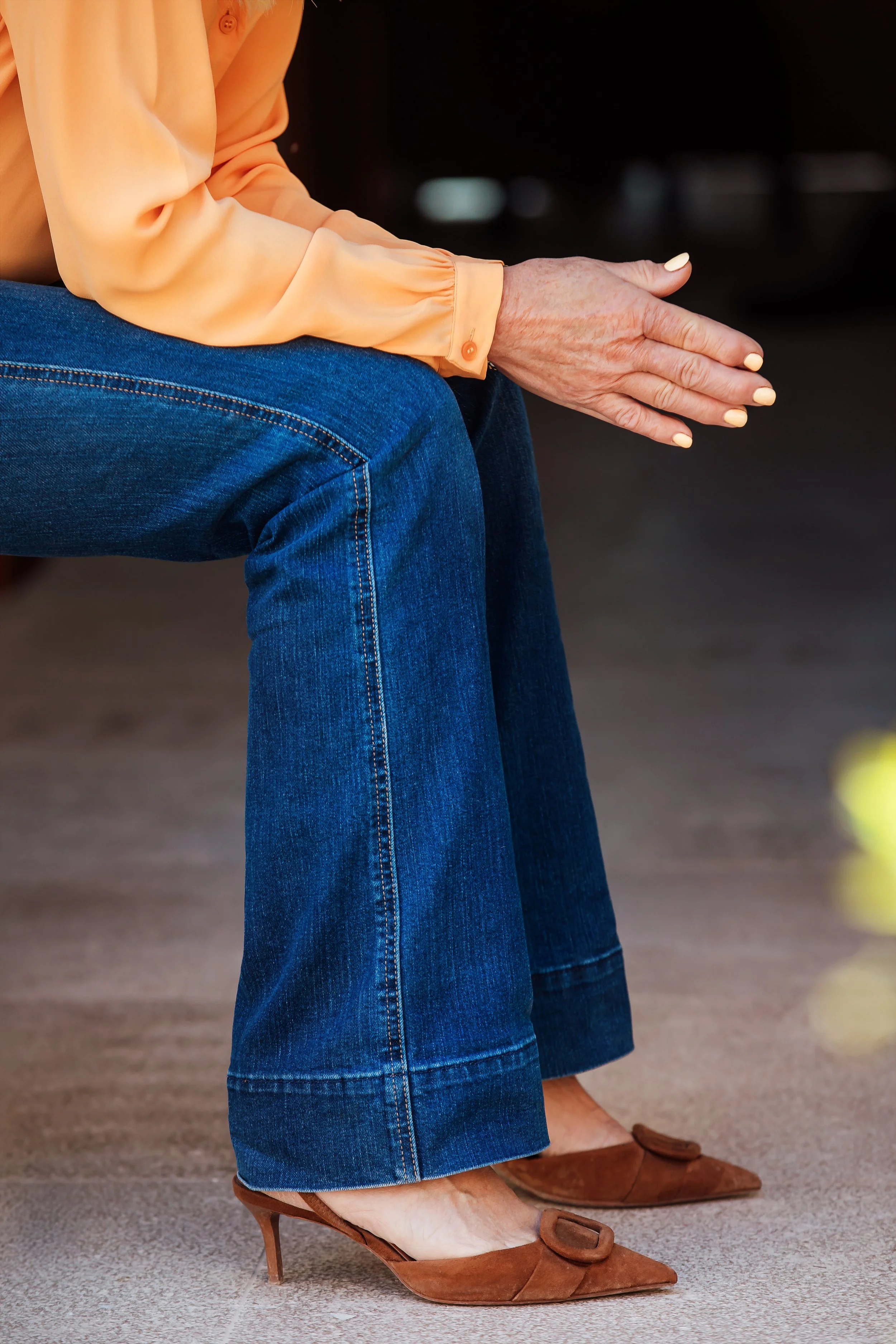 Närbild på en sittande person klädd i blå jeans, bruna högklackade skor och en persikofärgad blus.
