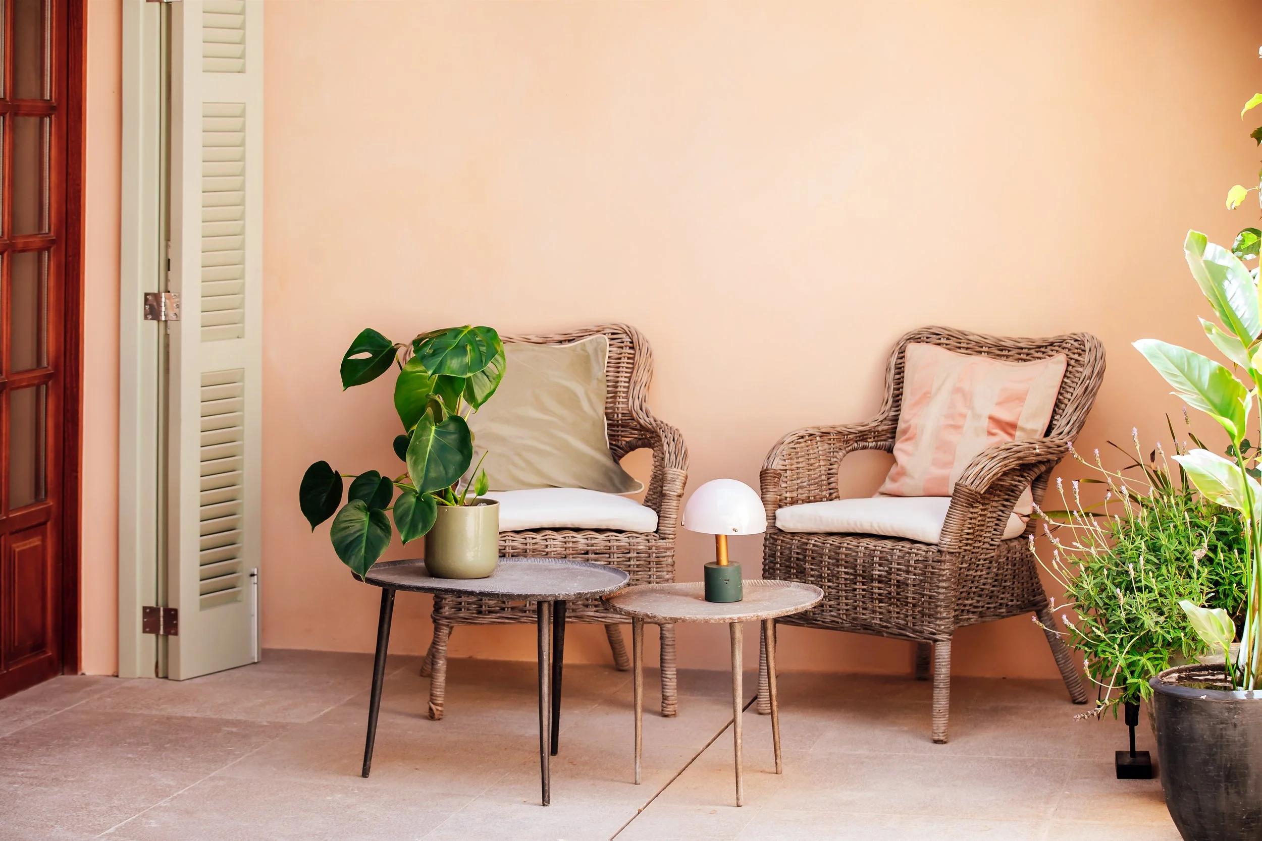 A cozy indoor sitting area with rattan furniture, including two chairs with beige cushions, two small patterned side tables, and various potted green plants against a peach-colored wall. A small white lamp is on one table, and a larger potted plant is near the door.