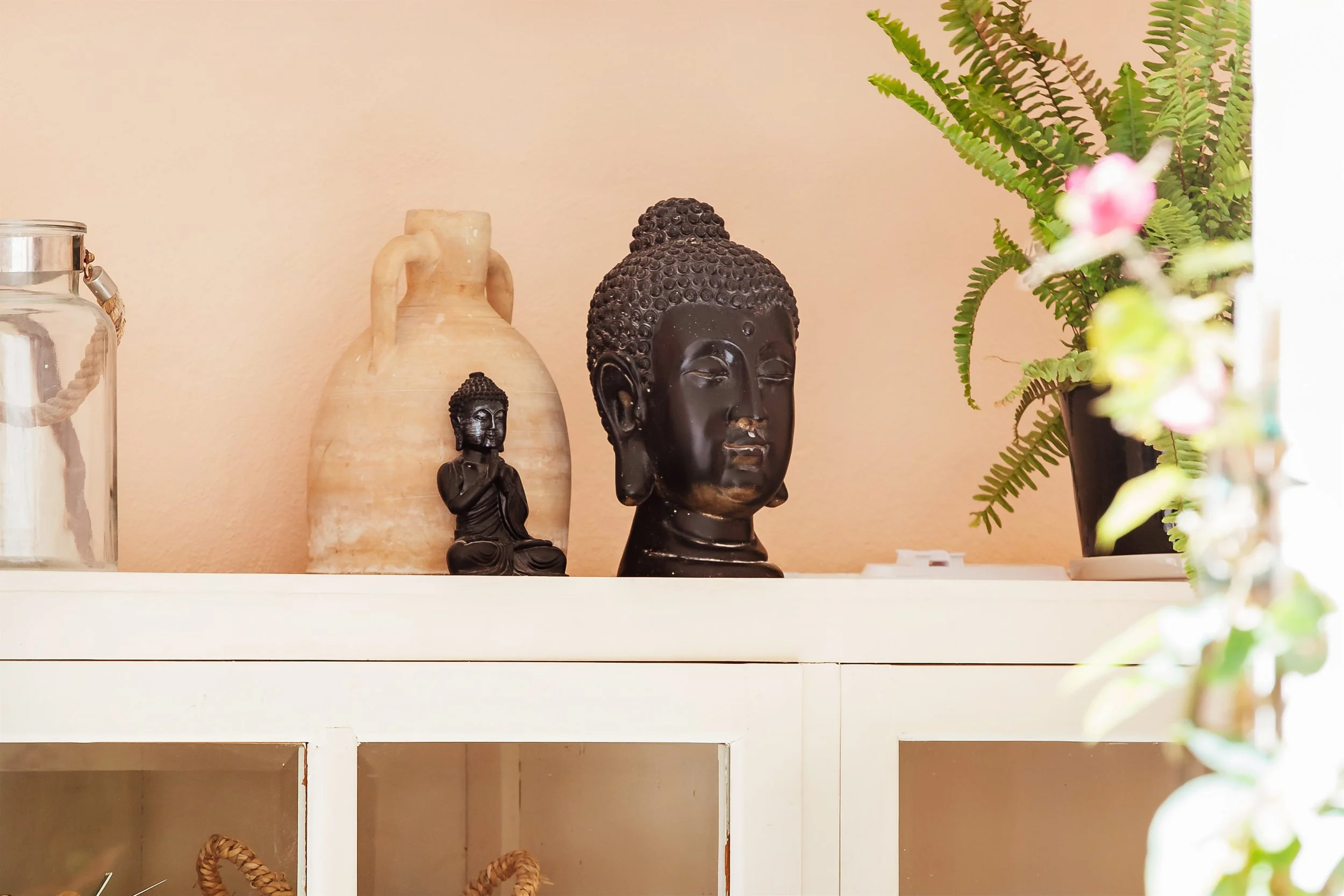 En hylla med olika dekorativa föremål, bland annat en stor keramikkanna, tre svarta Buddhastatyer i olika storlekar och en svart vas med gröna ormbunkar.