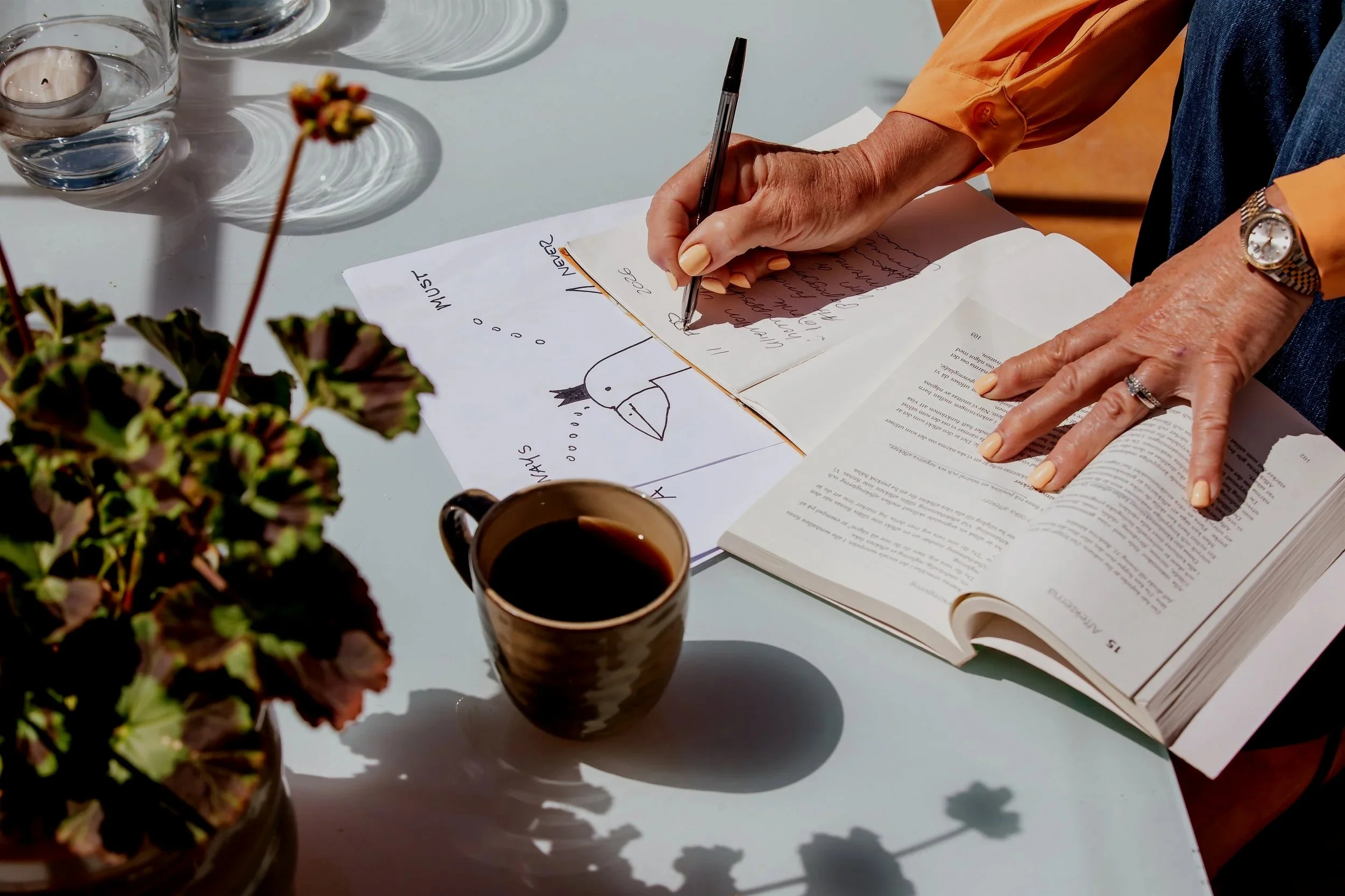 En person skriver anteckningar i en almanacka. På bilden syns en fågel omgiven av en bok, en kopp kaffe, en glasskål med ett ljus och krukväxter på ett vitt bord.