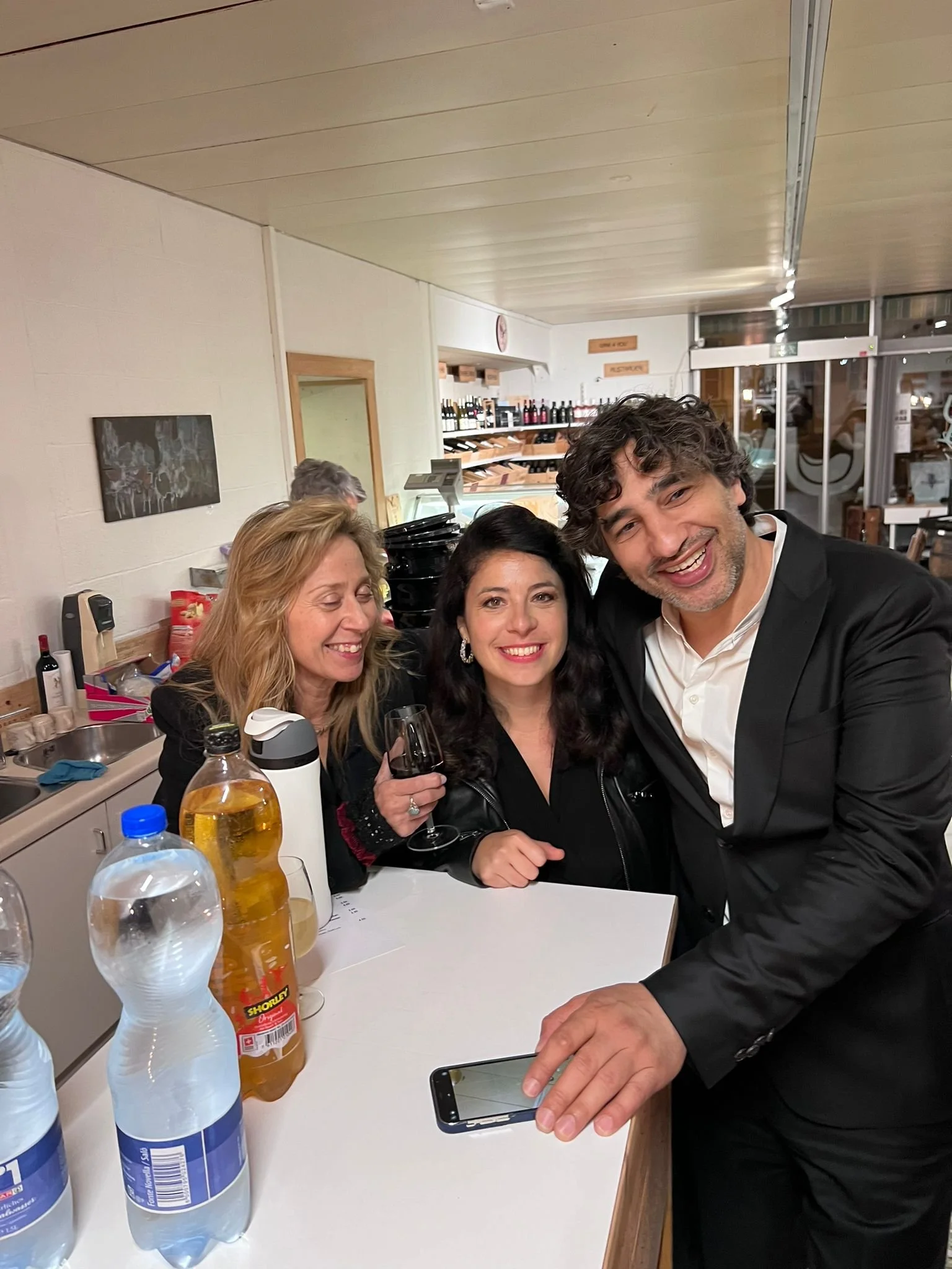 Three people smiling and posing for a photo in a restaurant or bar, with drinks and bottled water on the counter in front of them.