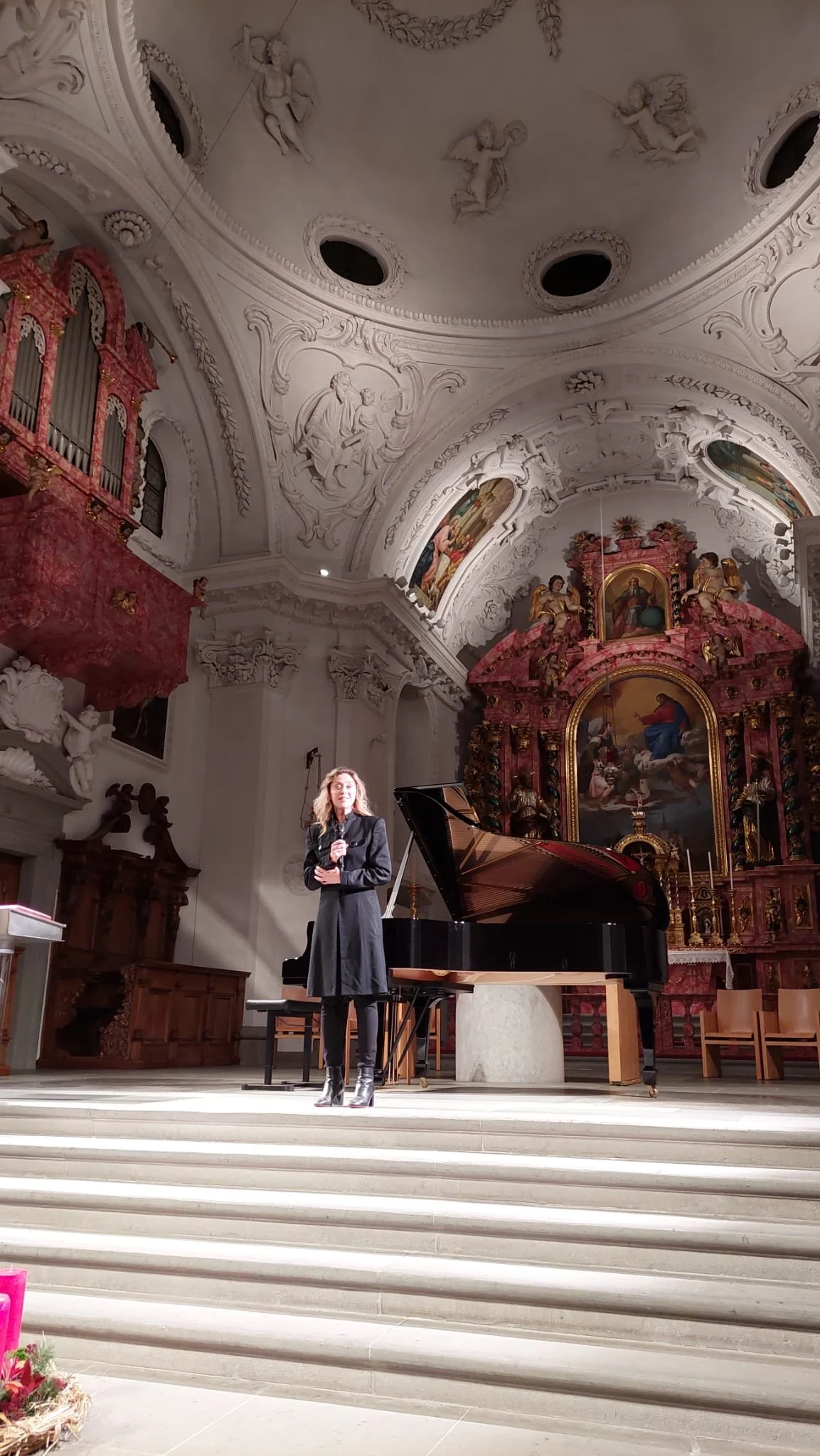 A woman standing on a church altar, holding a microphone, with a grand piano nearby and religious artwork and sculptures in the background.