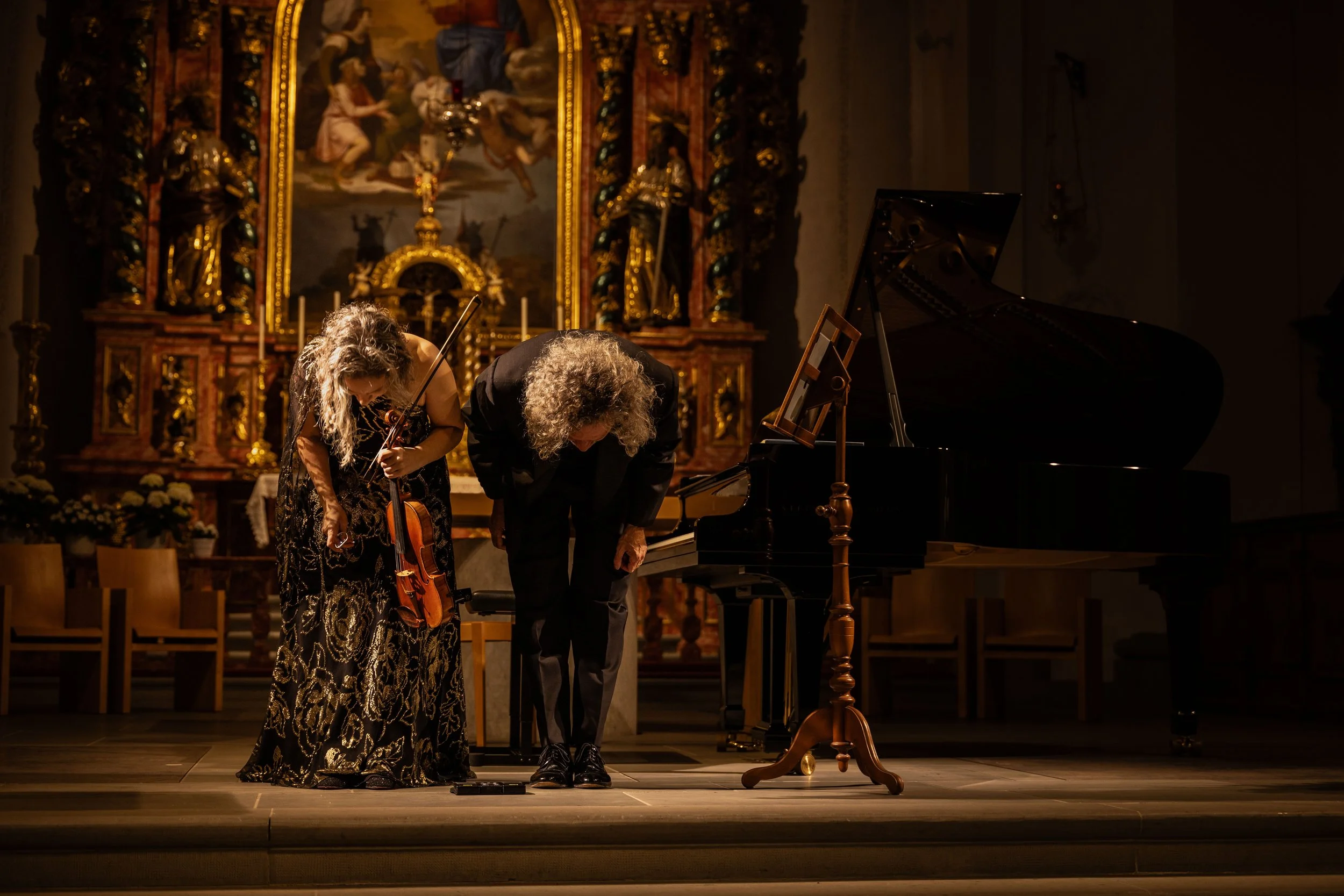 Two musicians, a woman holding a violin and a man bowing, stand on a stage with a grand piano before a religious altar in a church. The woman is dressed in a black and gold patterned gown, and the man in a black suit. The altar behind them features a