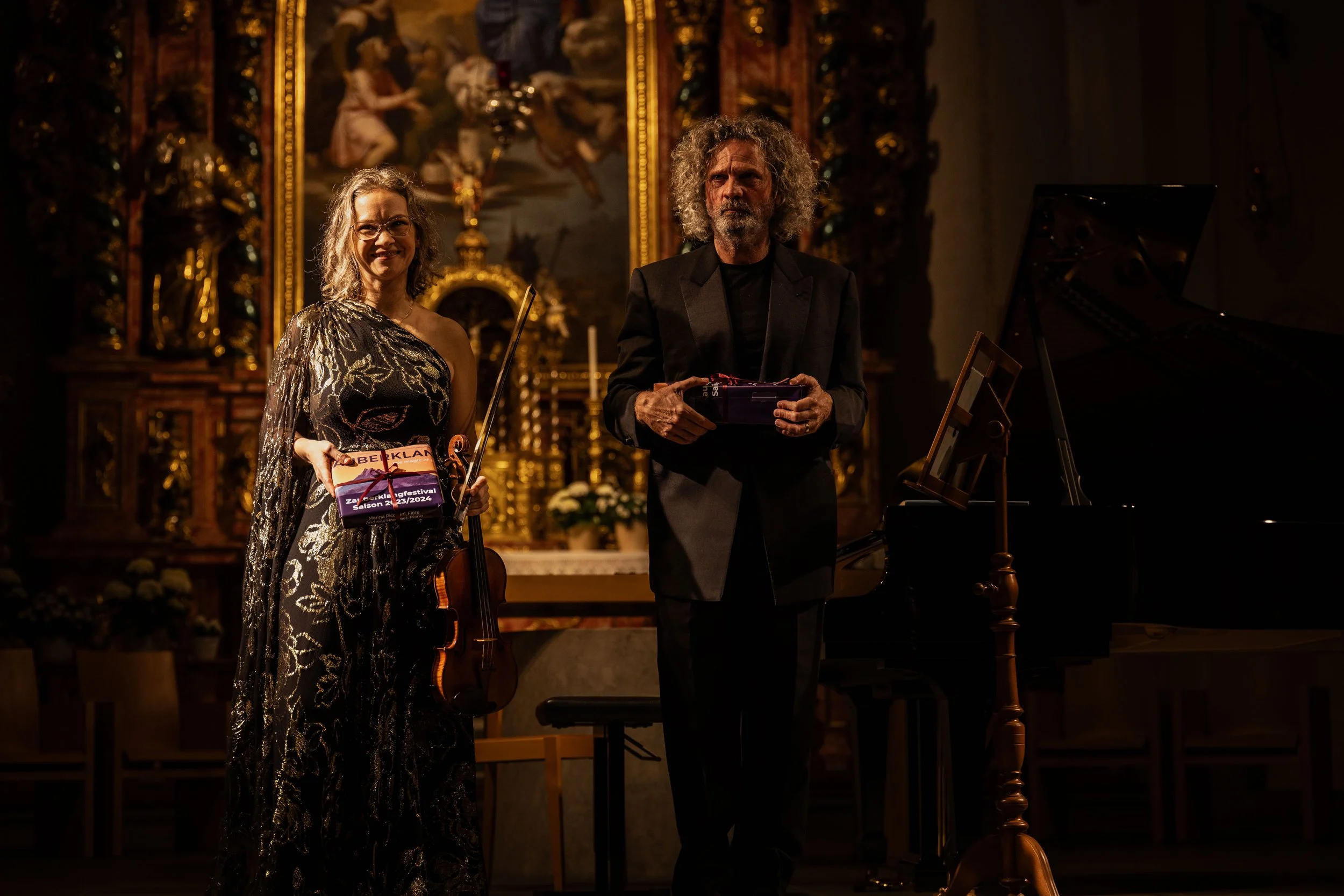 A woman holding a violin and a box standing next to a man holding a box, both on stage with a grand piano and ornate church background.