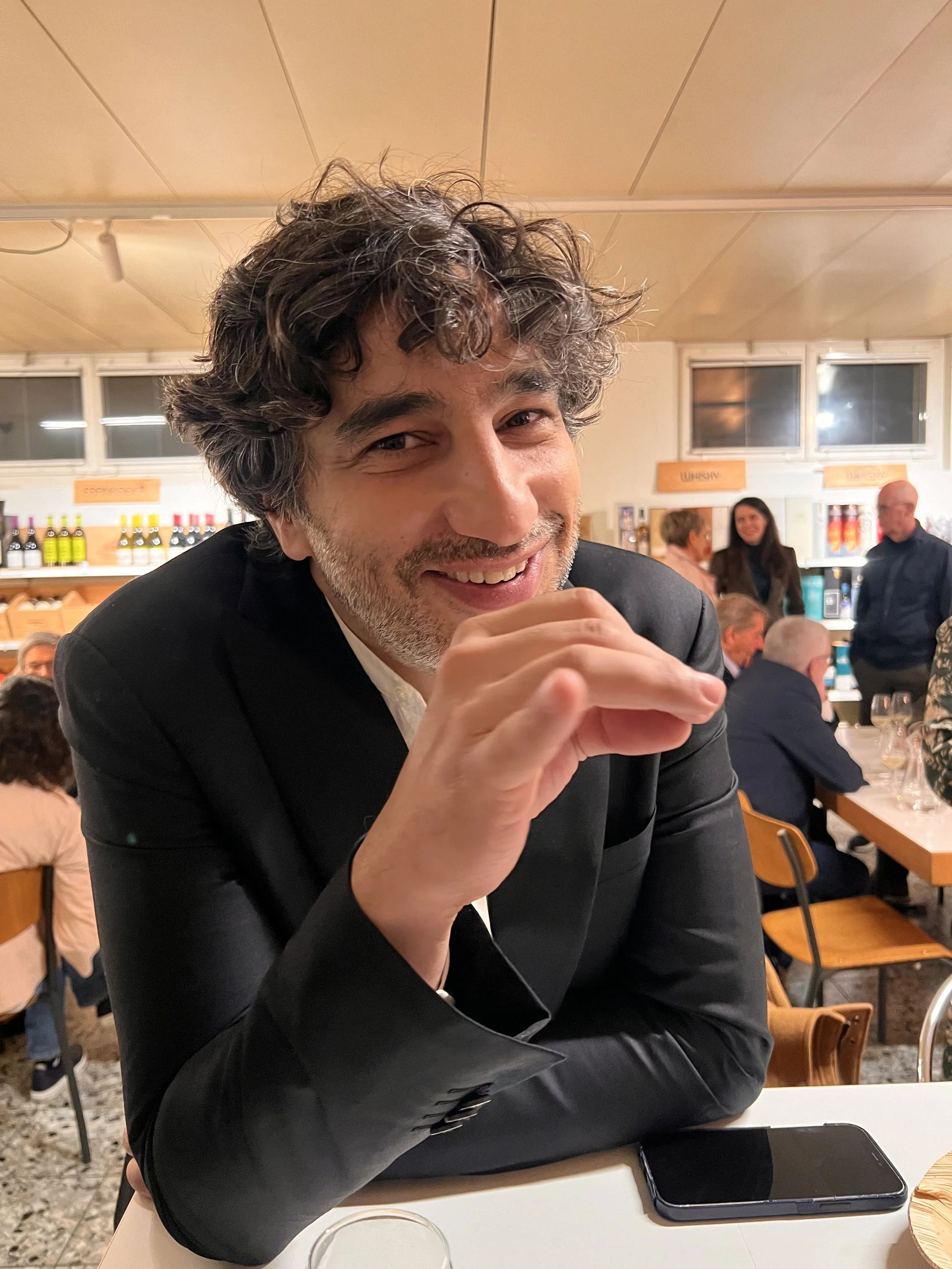 A man with curly hair and a beard smiling at the camera while seated at a table in a busy restaurant or bar with other people in the background.