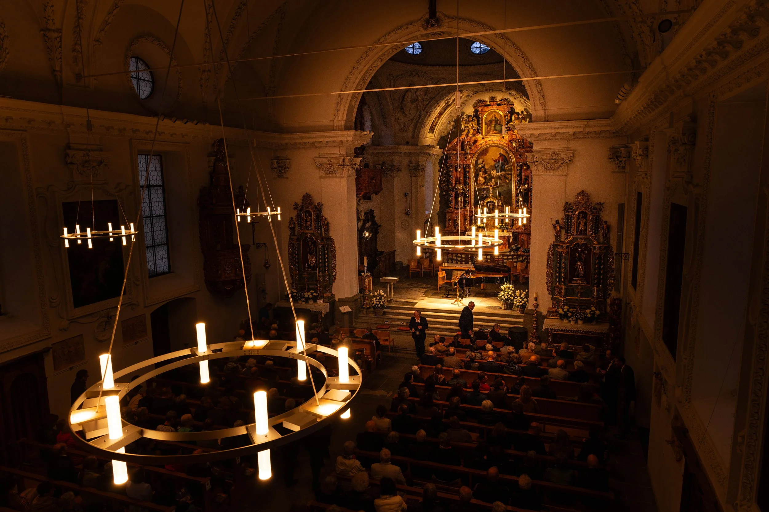 Interior of a church during a performance, with audience seated, candles, and ornate altar in the background.