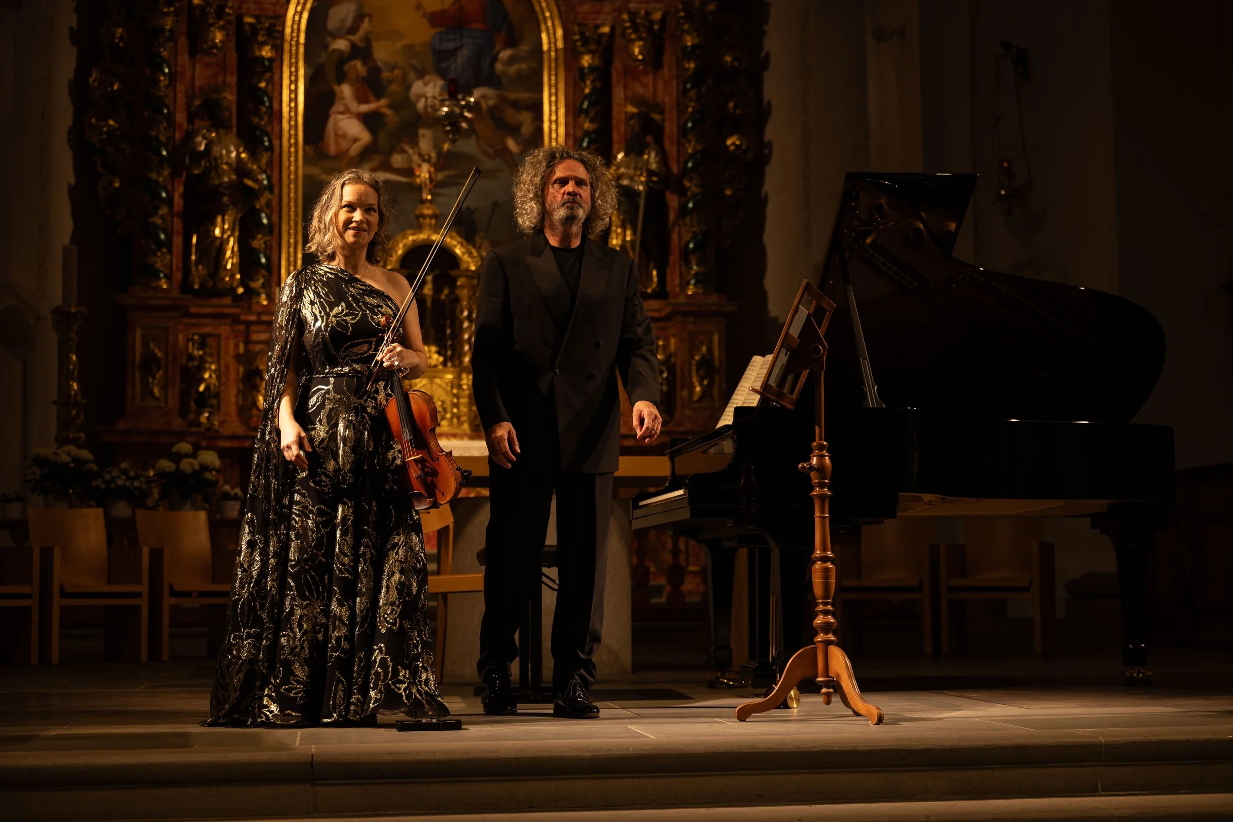 Two musicians stand on stage in a church or concert hall, one woman holding a violin and bow, the man standing beside a grand piano. They are dressed formally, and the background features an ornate altar or altarpiece with religious artwork and gold