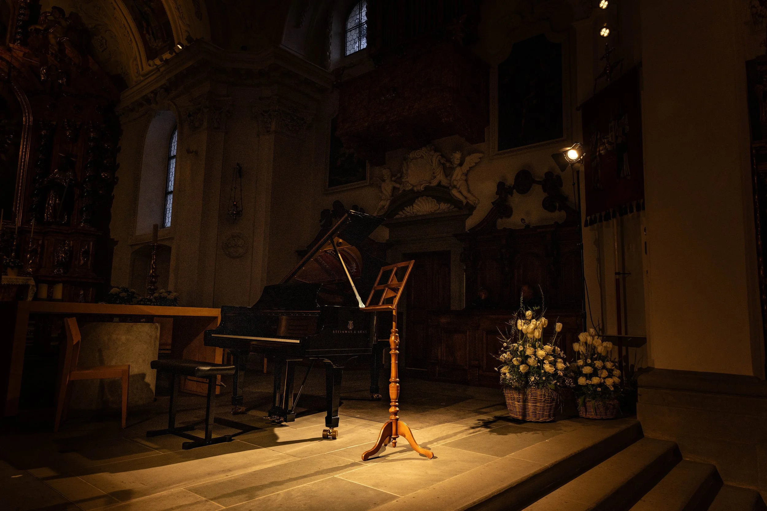 Empty grand piano on a small stage in a dimly lit church, with a music stand and flower arrangements nearby.