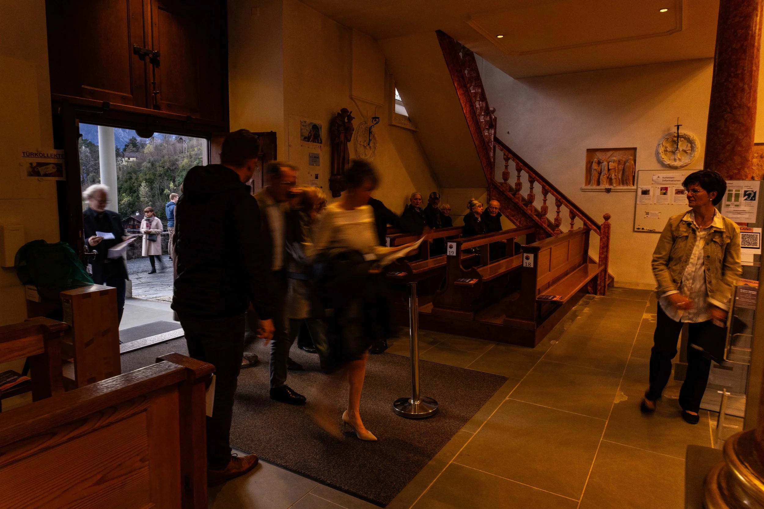People entering a church or hall through an open door, with some sitting inside and others standing outside, in a cozy, wooden interior with religious decorations.