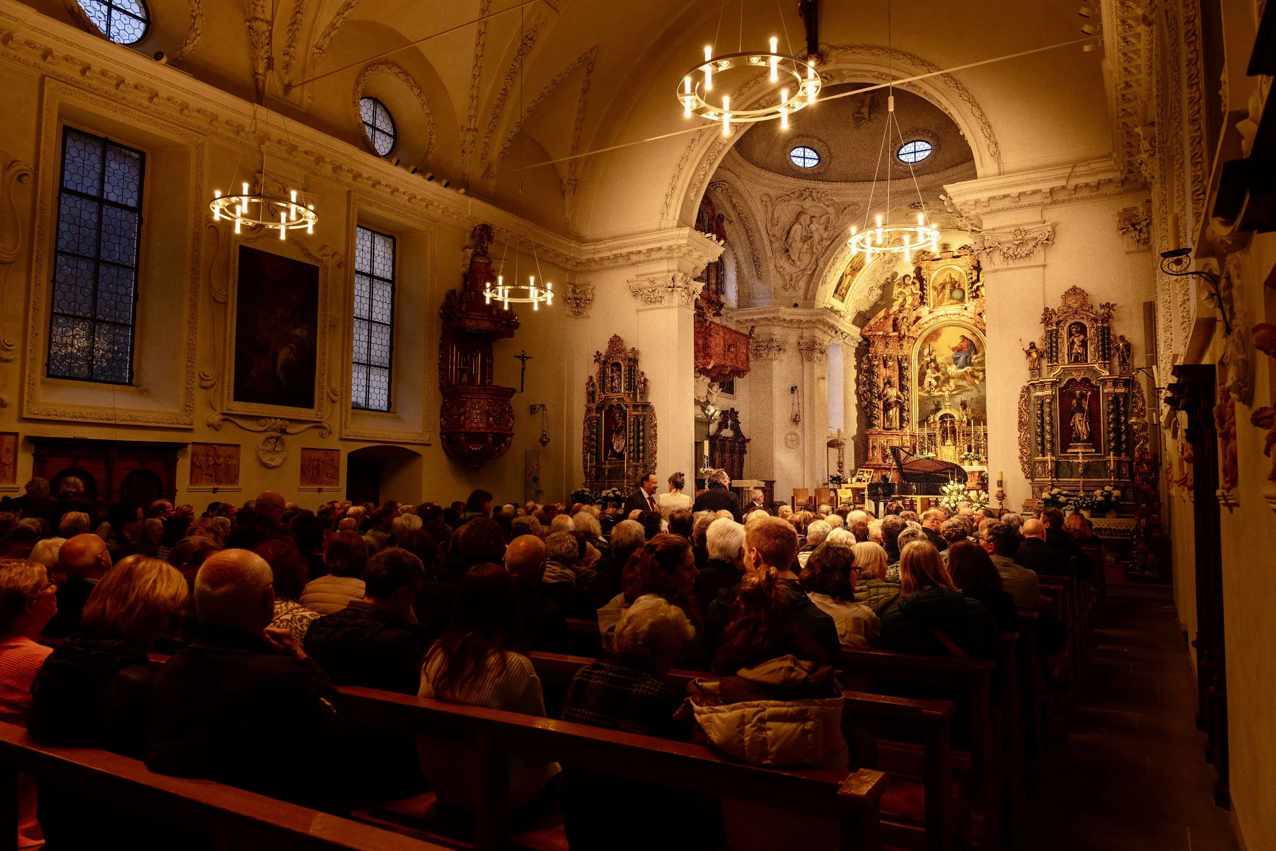 Inside a church, a large congregation seated on wooden pews, facing an ornate altar with religious artwork and sculptures, illuminated by chandeliers hanging from a high, decorated ceiling.