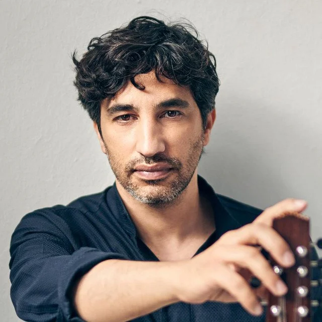 Man with dark curly hair and stubble, wearing a dark shirt, holding a guitar.