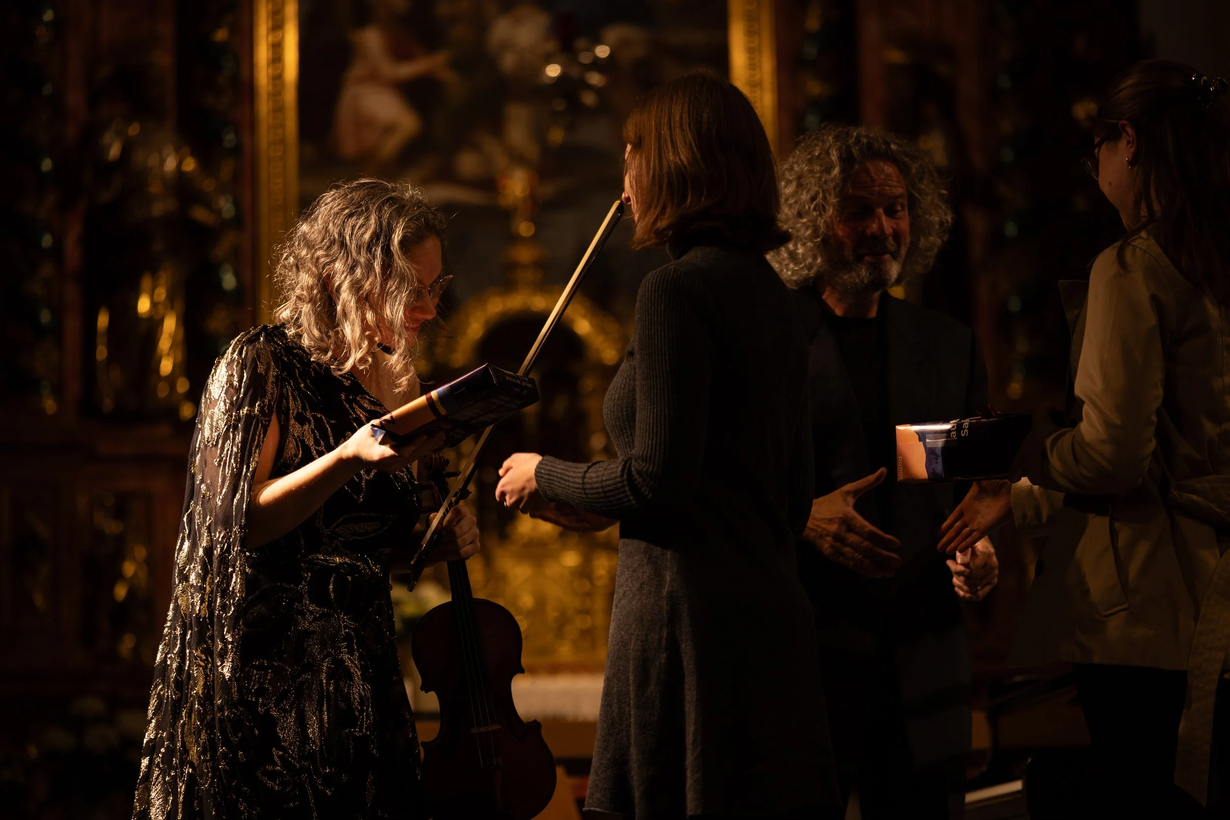 Four people engaged in a conversation indoors near a gold ornate background, one woman with curly hair holding a map or booklet and a violin, another woman with straight hair, man with curly hair and beard, and a woman with glasses, all in a dimly li