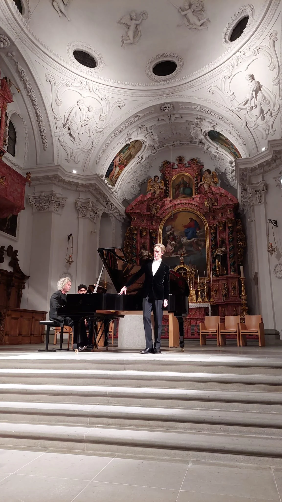 A musical performance inside a grand, ornate church with a white ceiling adorned with detailed sculptures and paintings. A woman in a black suit is singing at a grand piano, accompanied by a pianist and another musician in the background.