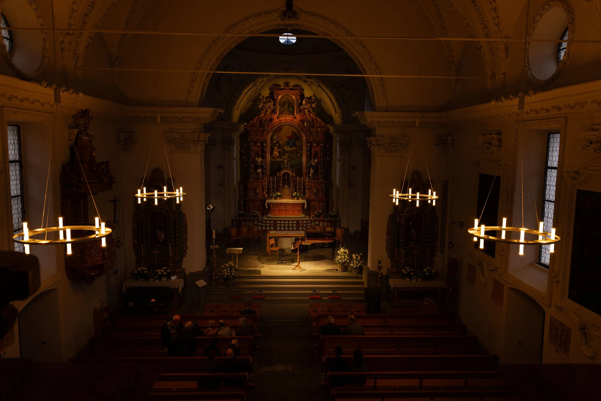 Interior of a church with ornate altar, wooden benches, and evening lighting from chandeliers.