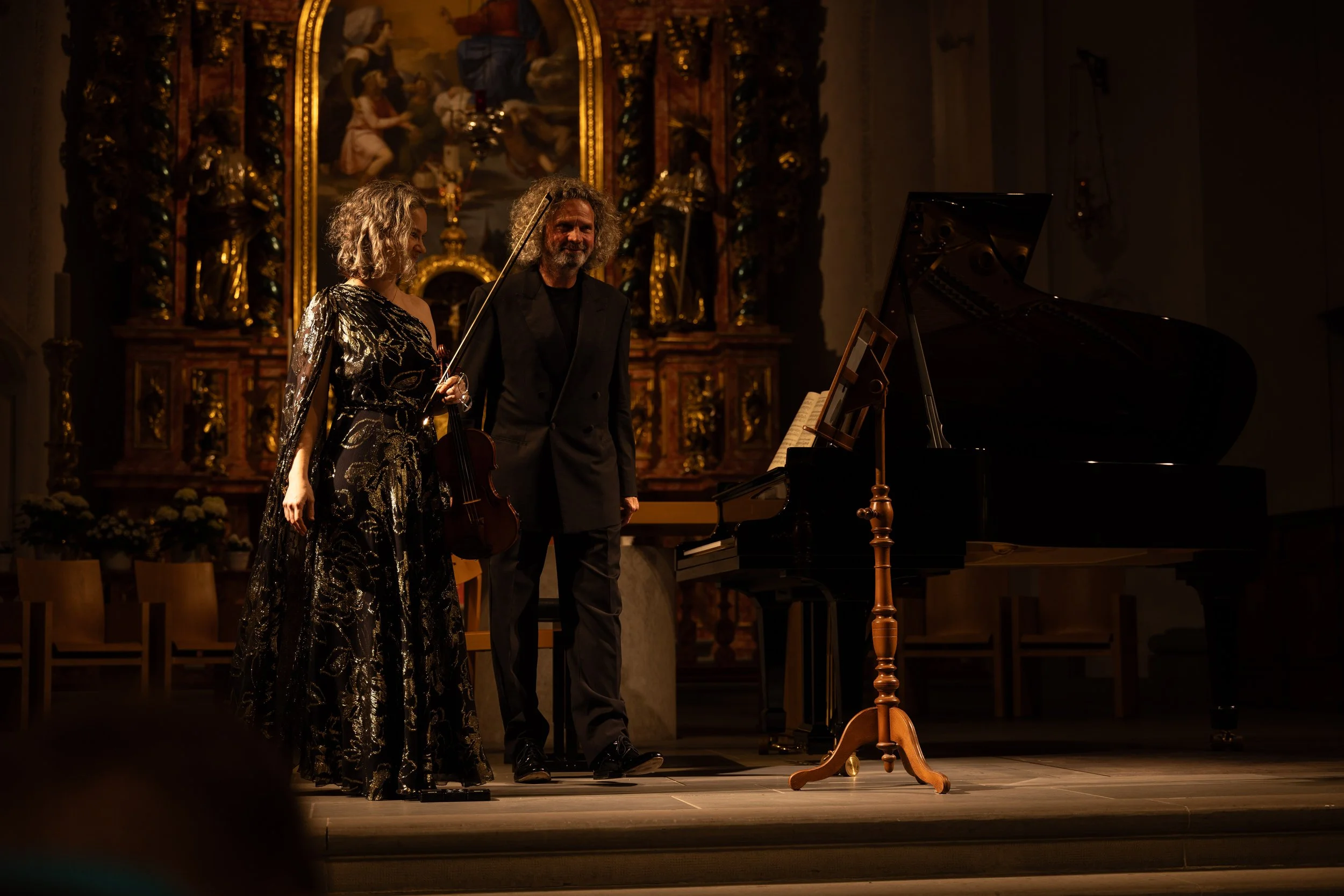 Two classical musicians, a woman holding a violin and a man with a bow, standing on stage after a performance, with a grand piano and ornate religious art in the background.